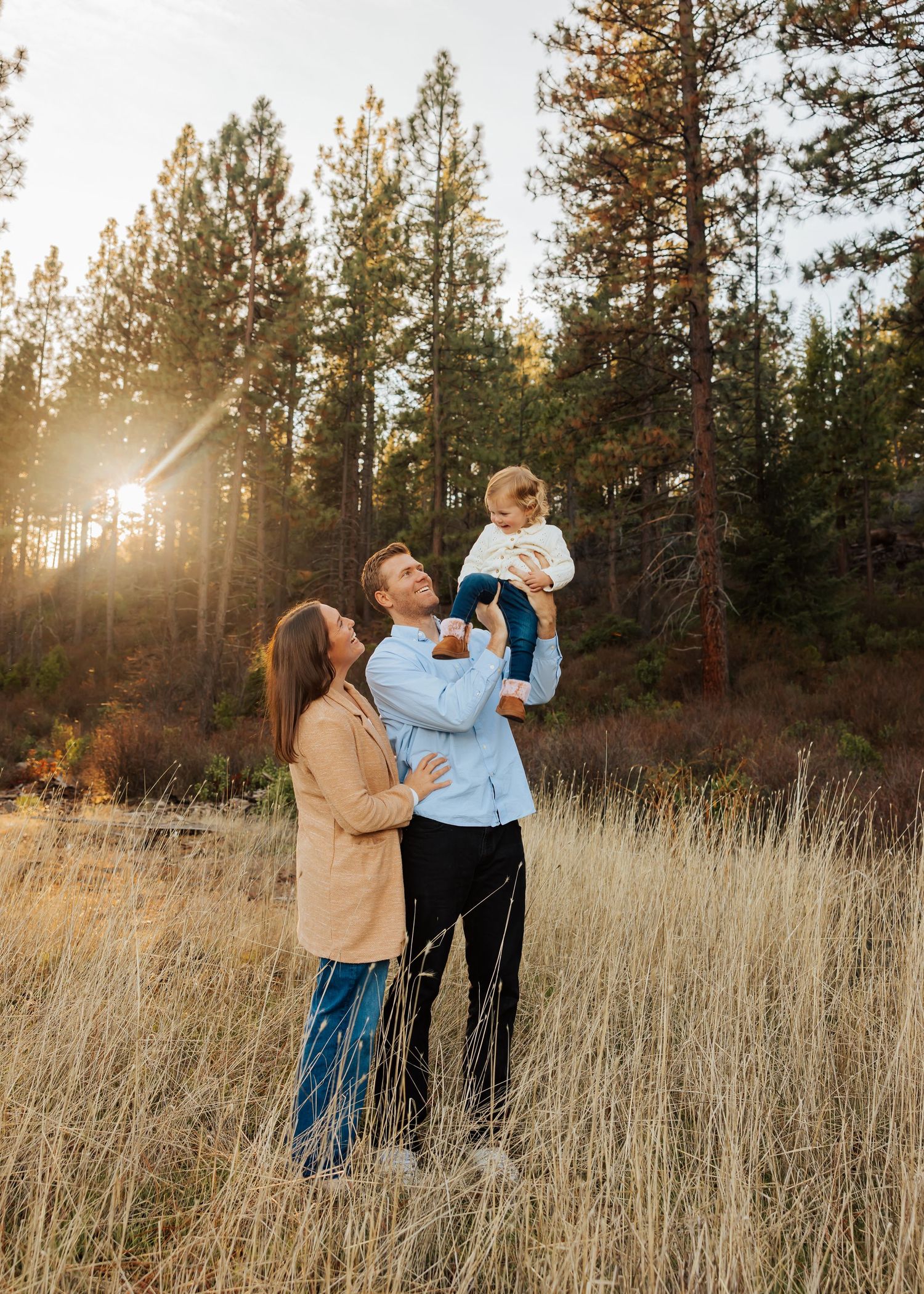 Joyful family moment captured in golden field with sunlit pine forest background.