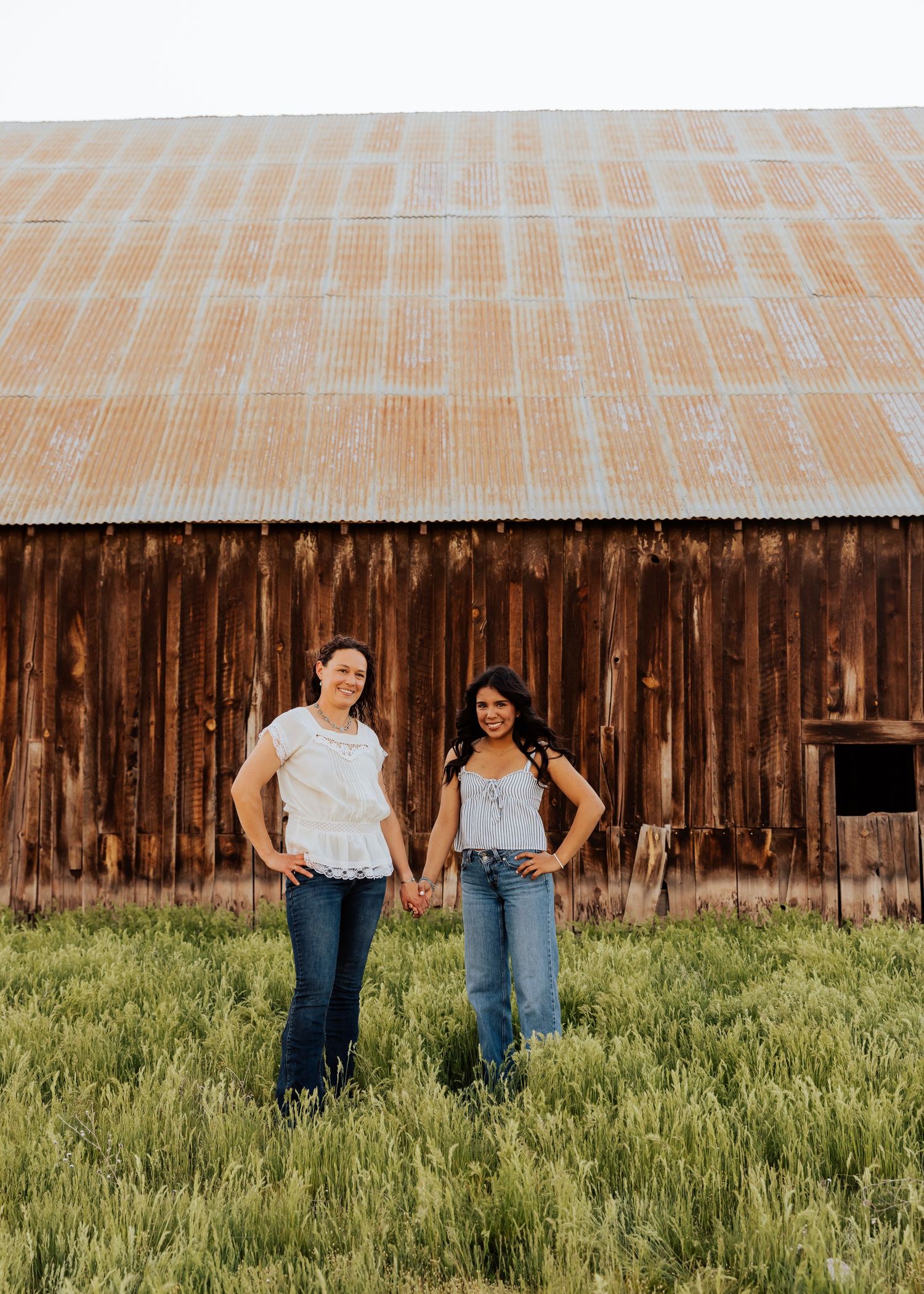 Two people stand together in casual attire against wooden barn backdrop and green field.