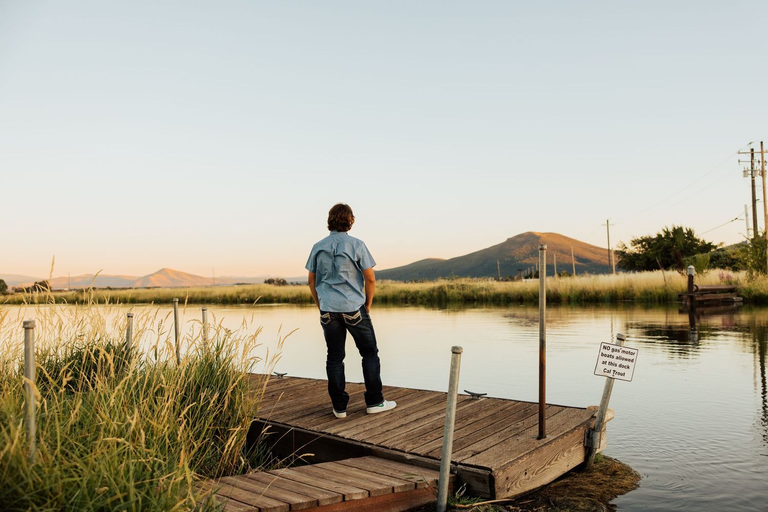 A distant figure stands on a wooden pier overlooking a serene lake at sunset.