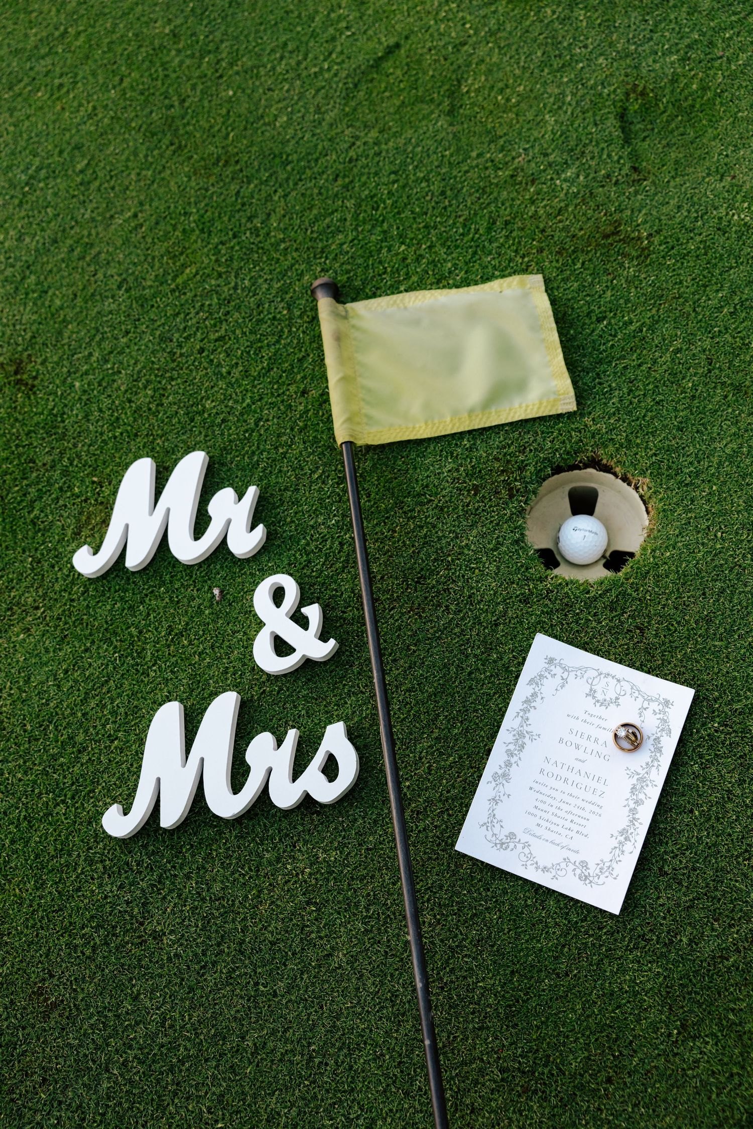 Mr & Mrs signs with golf flag, ball in hole, and wedding invitation on green grass course.