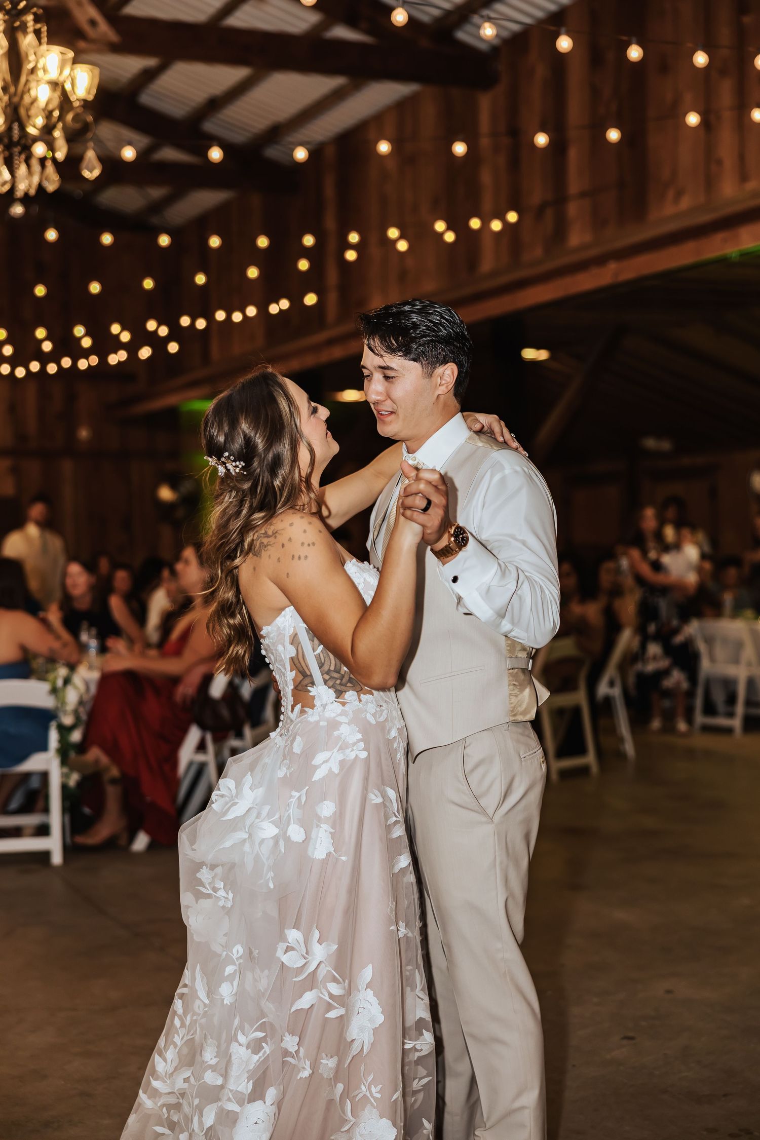 Bride and groom share their first dance in a rustic barn wedding venue with string lights glowing overhead.