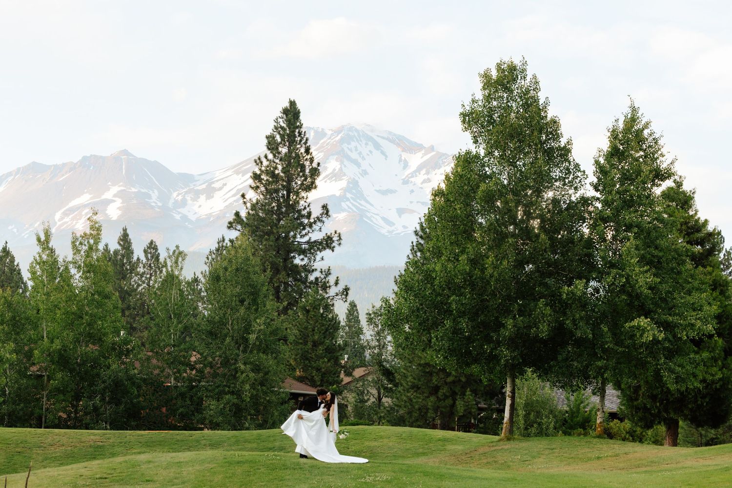 Bride and groom walk across lush green lawn with tall trees and snow-capped mountain backdrop at outdoor wedding.