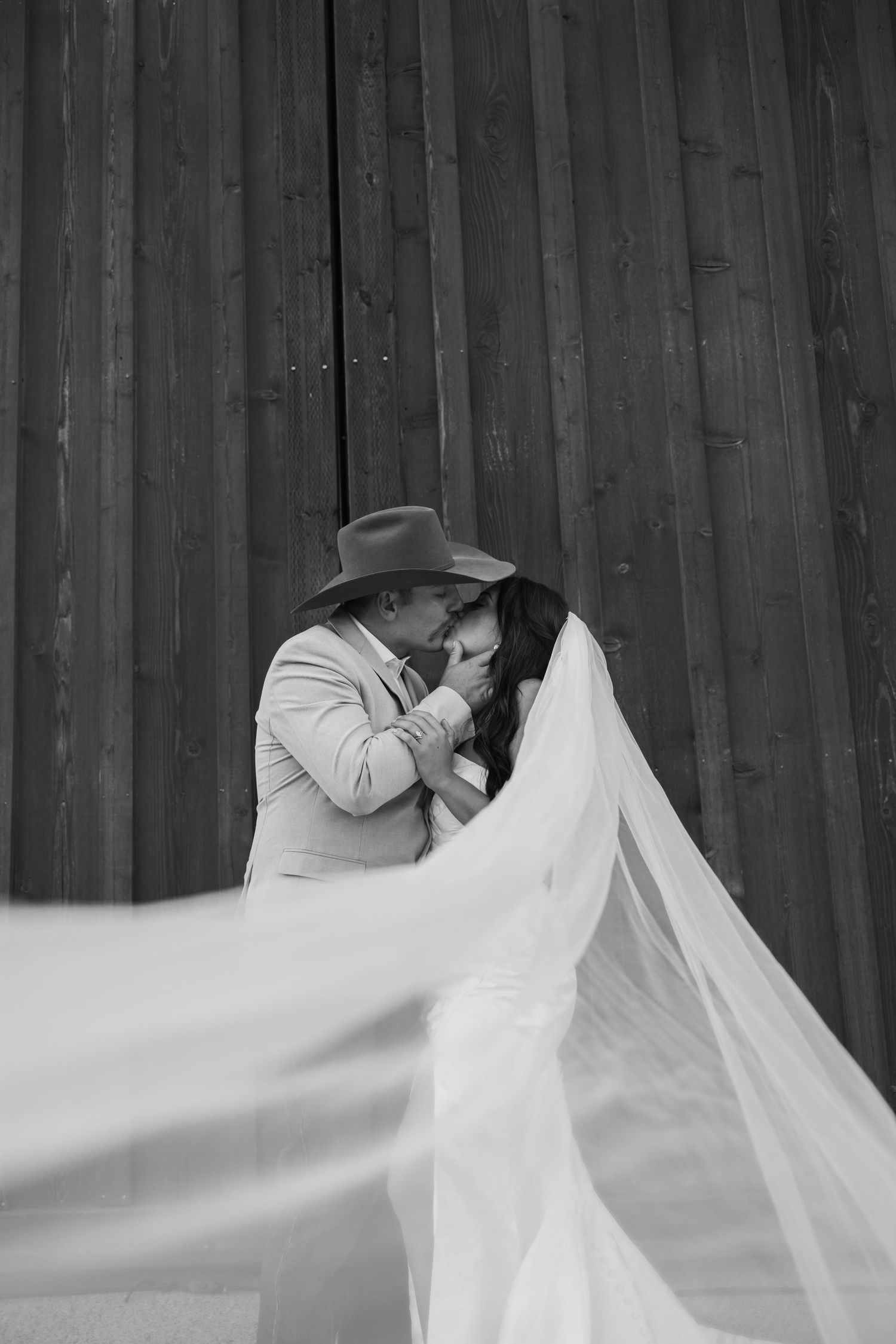 Artistic black and white wedding portrait with flowing veil against wooden barn wall.