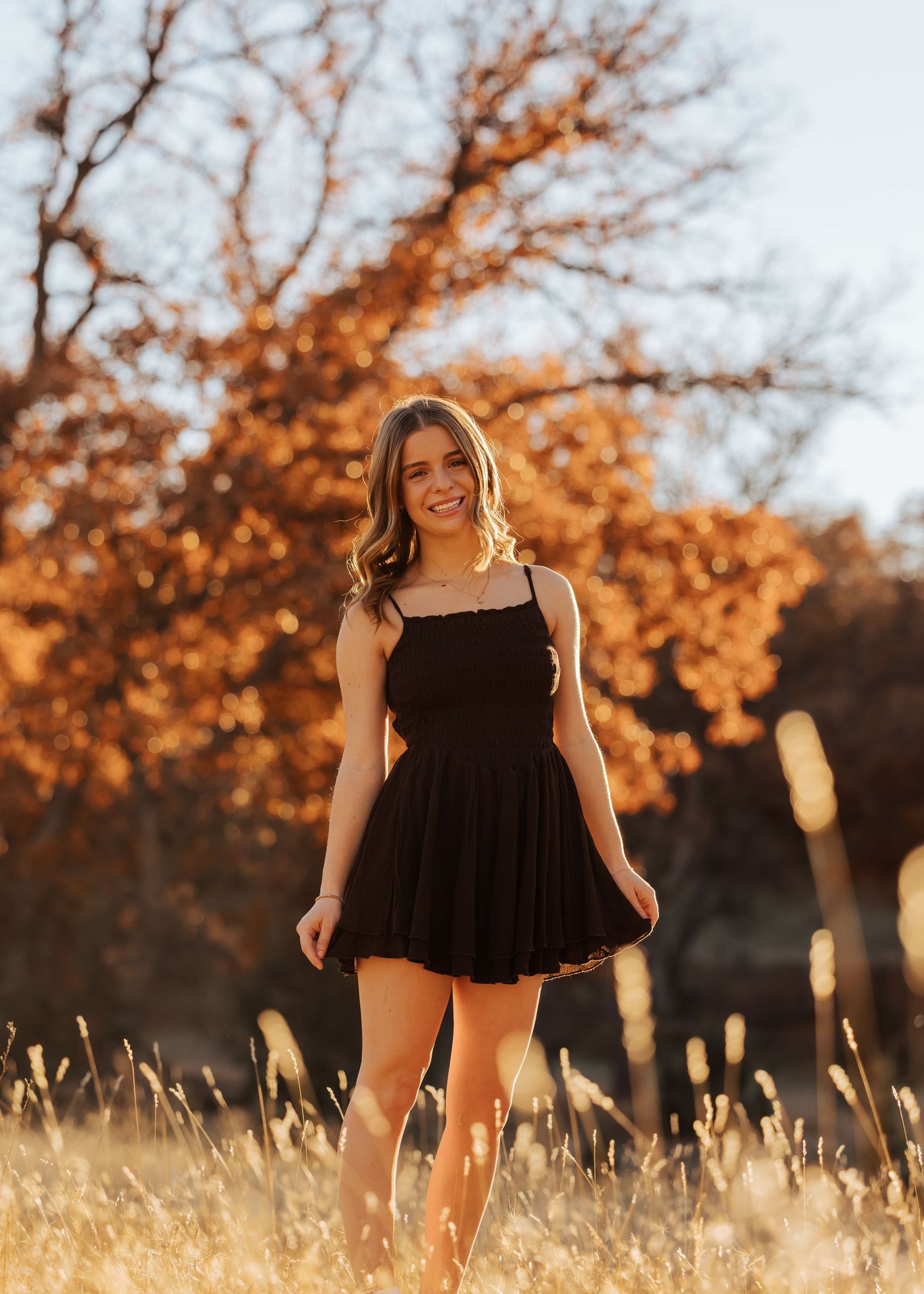 Someone in a black dress poses playfully in a sunlit field with autumn trees.