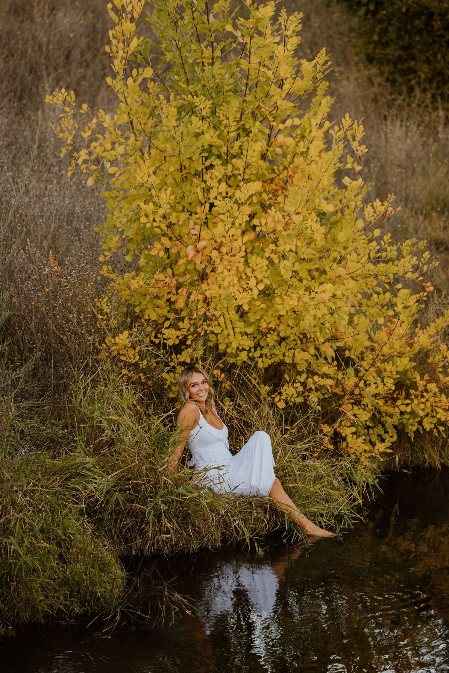 Woman in white dress sits by yellow autumn tree reflecting in calm water.