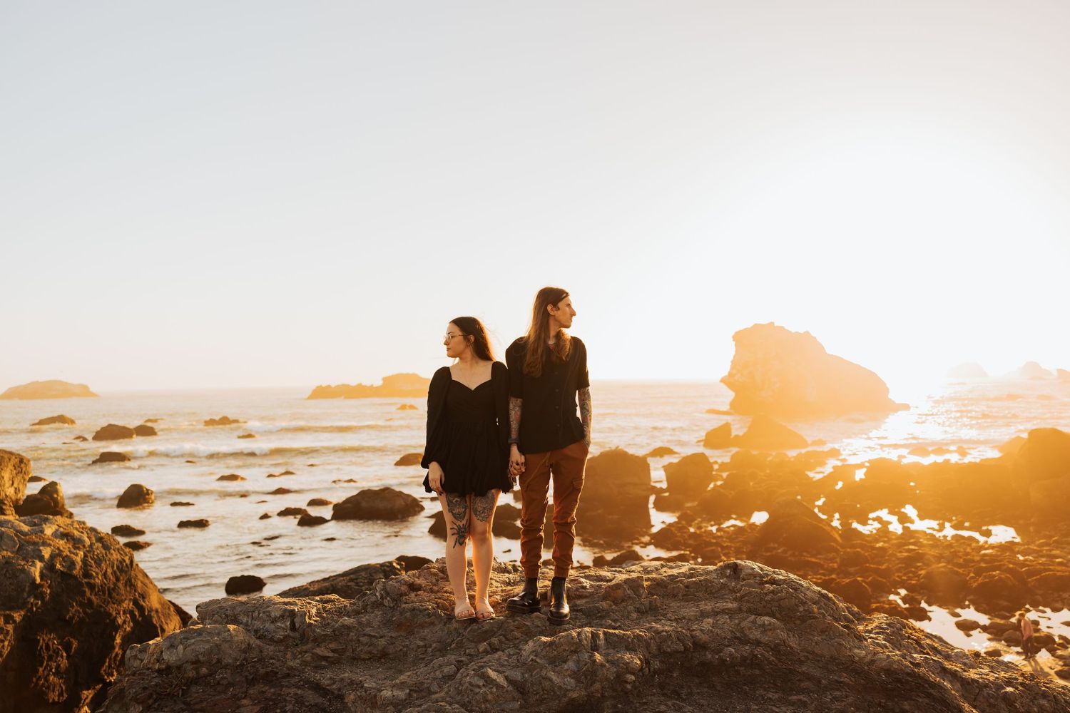 Two figures stand on coastal rocks at sunset with ocean waves in the background.
