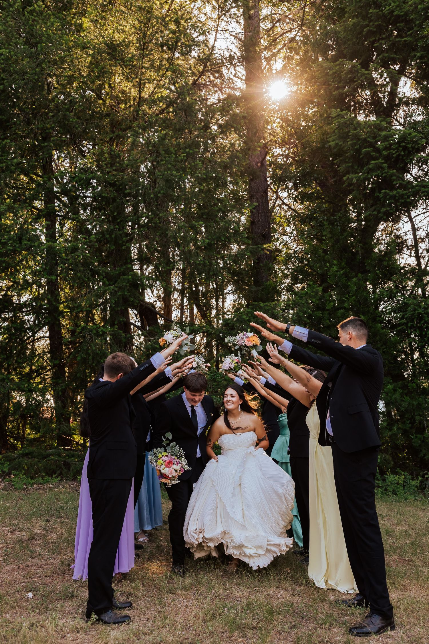 Wedding party forms an arch with arms as bride and groom walk through, surrounded by tall trees with golden sunlight.