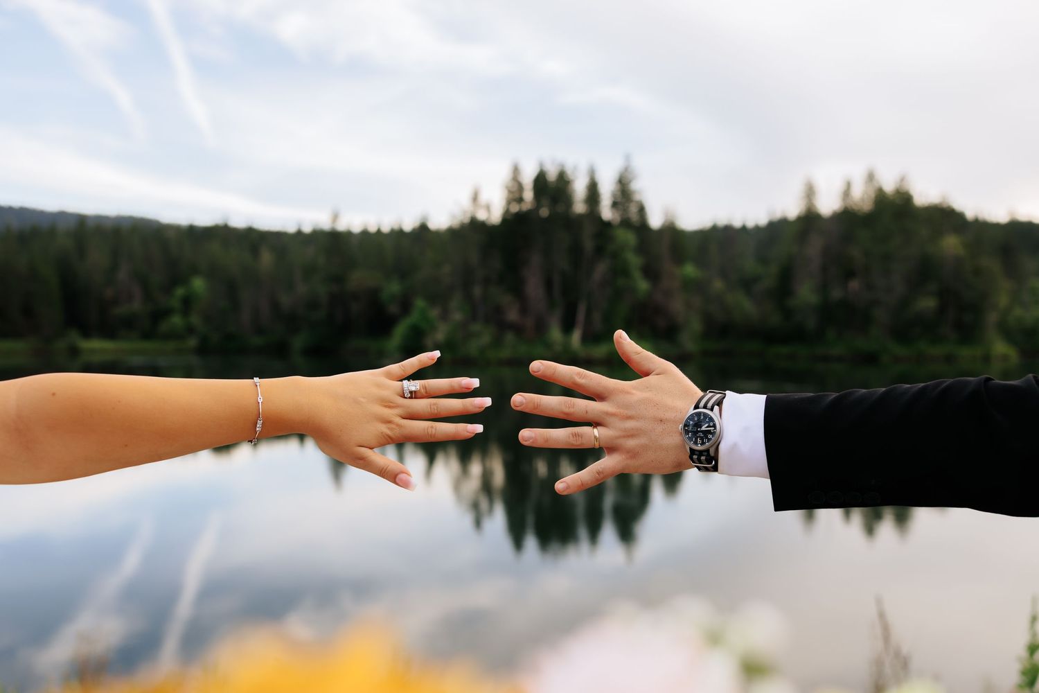Two hands reaching toward each other over a reflective lake, one wearing a wedding ring and bracelet, the other a watch.