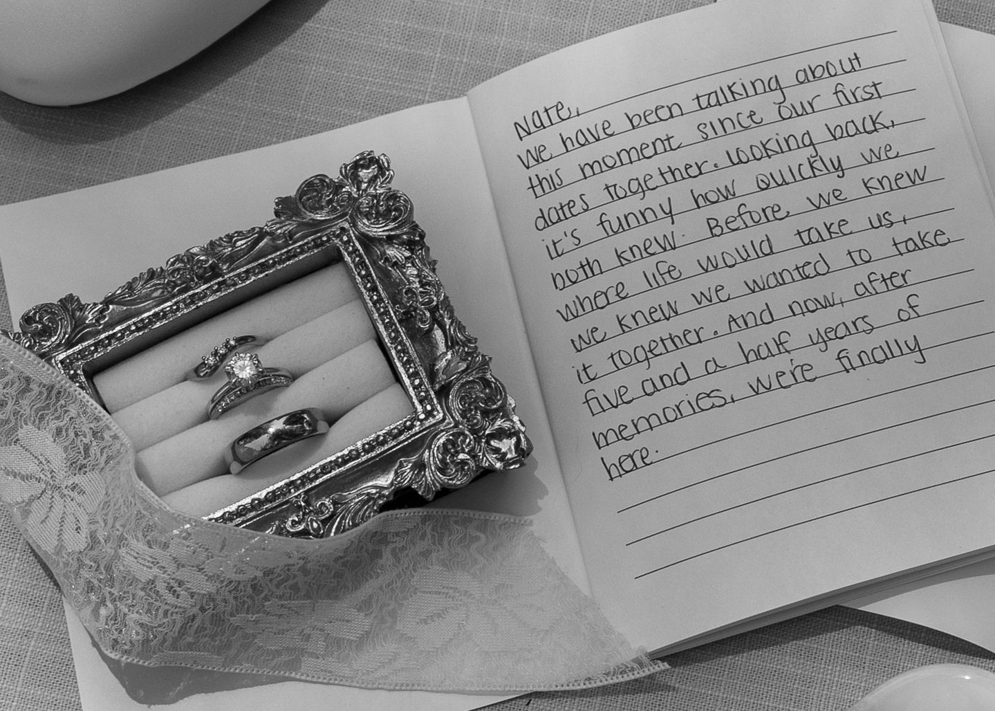 Black and white photo of engagement rings in ornate frame beside open journal with handwritten love note.