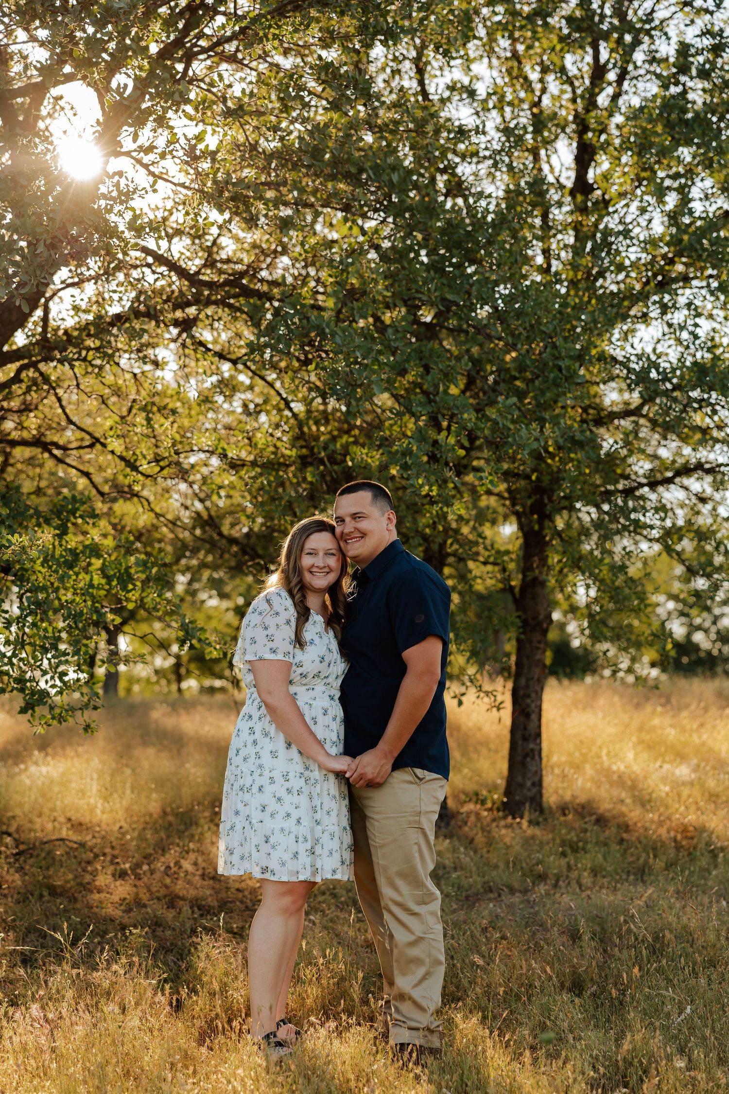 Couple smiling together in golden field at sunset with green trees behind them.