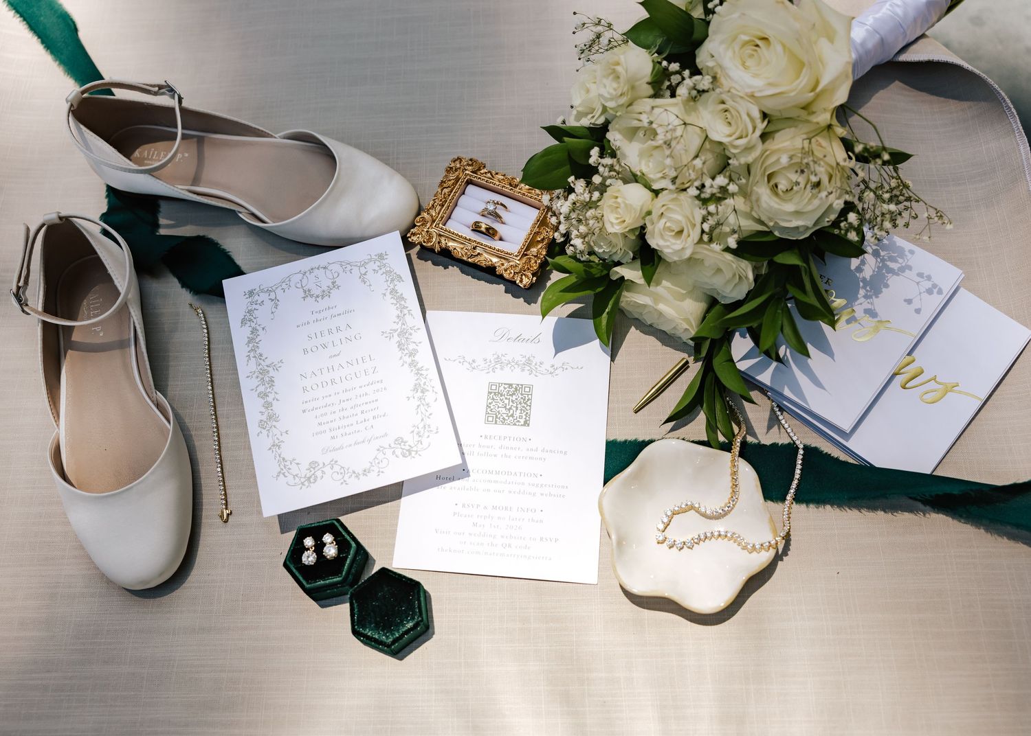 Elegant wedding flat lay with white heels, floral bouquet, invitations, pearl necklace, and green velvet ring box.
