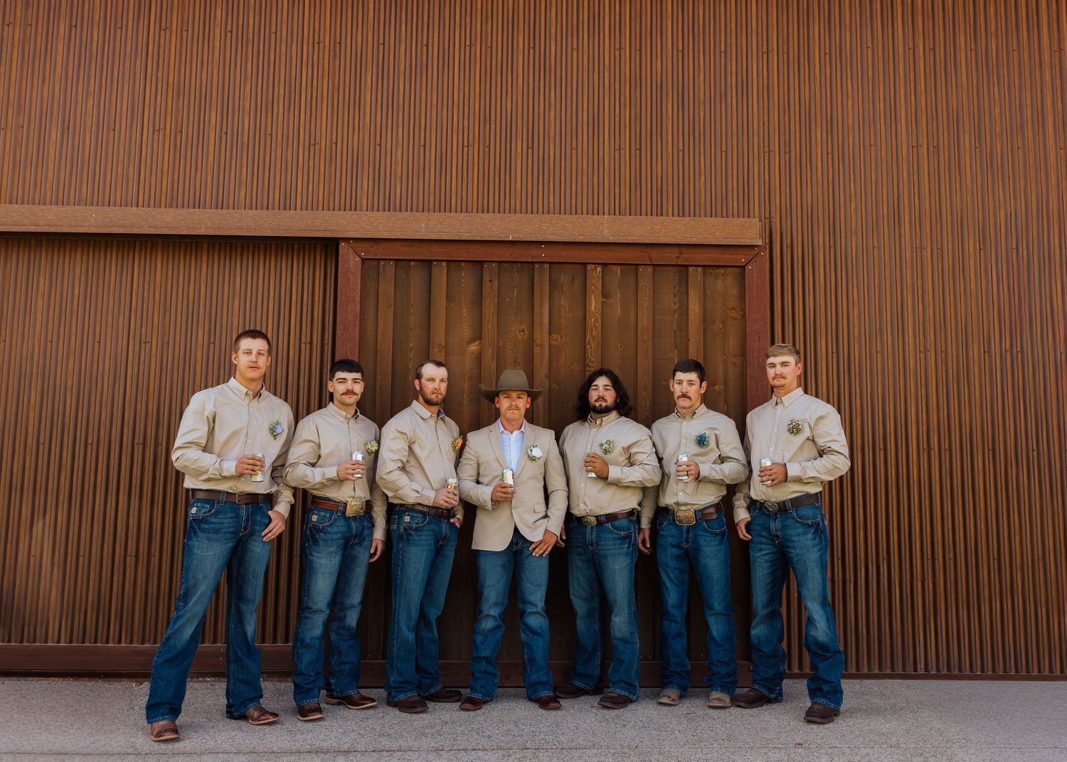 Groomsmen in matching outfits sit casually against a brown wall showing their boots.