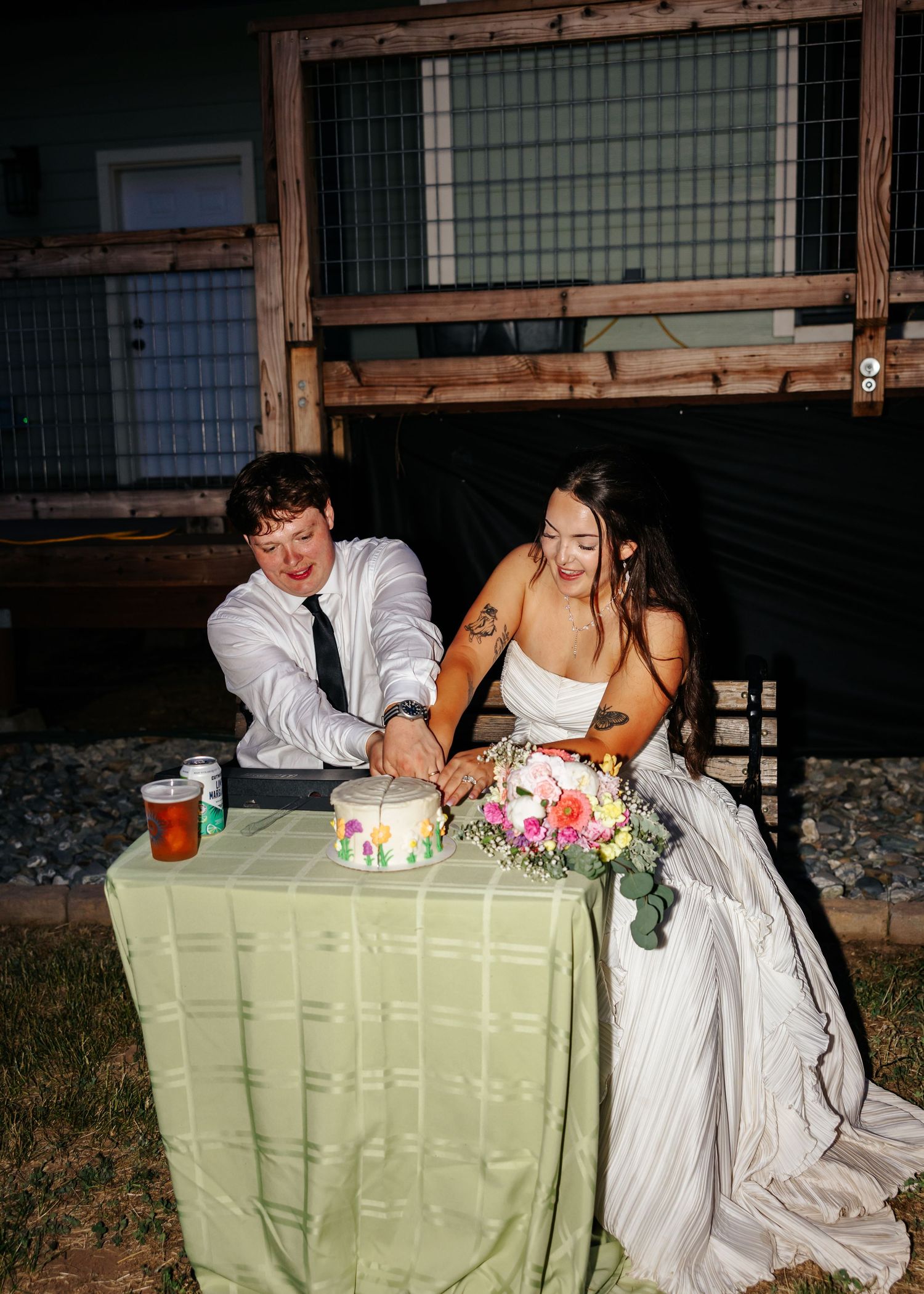 Bride and groom cutting wedding cake together at night outdoor reception, laughing joyfully.