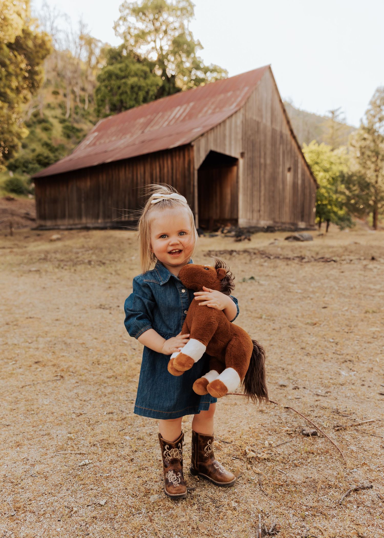 Child plays with toy horse in front of rustic barns on dry grass field.