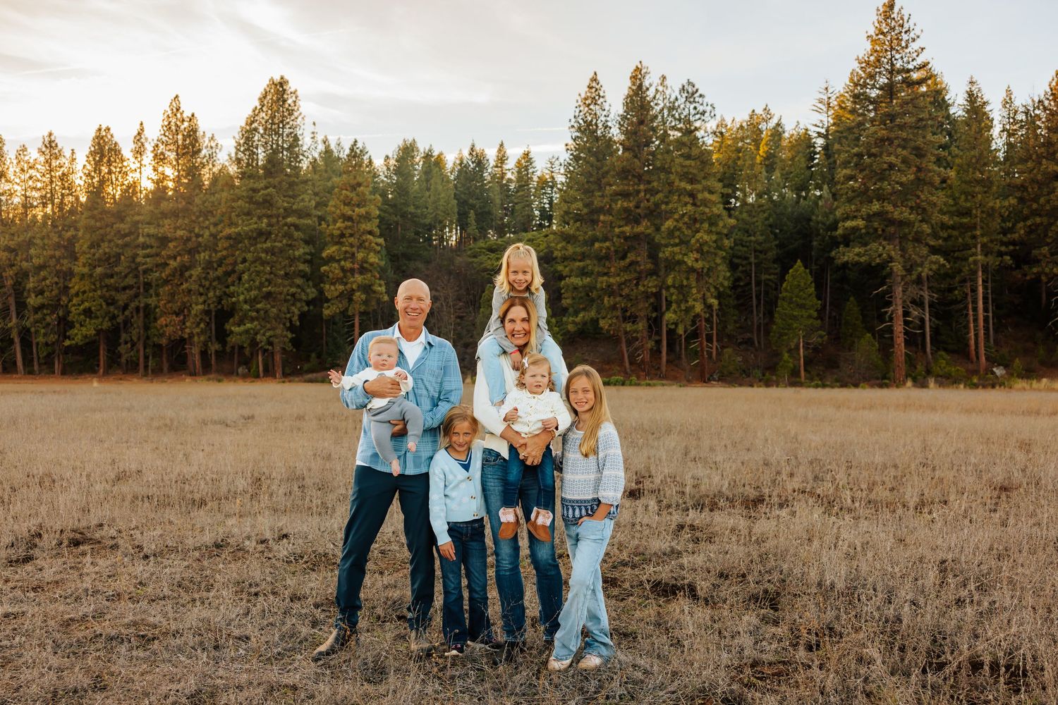 Small family group in coordinated blue and white outfits poses in an open field with pine forest backdrop.