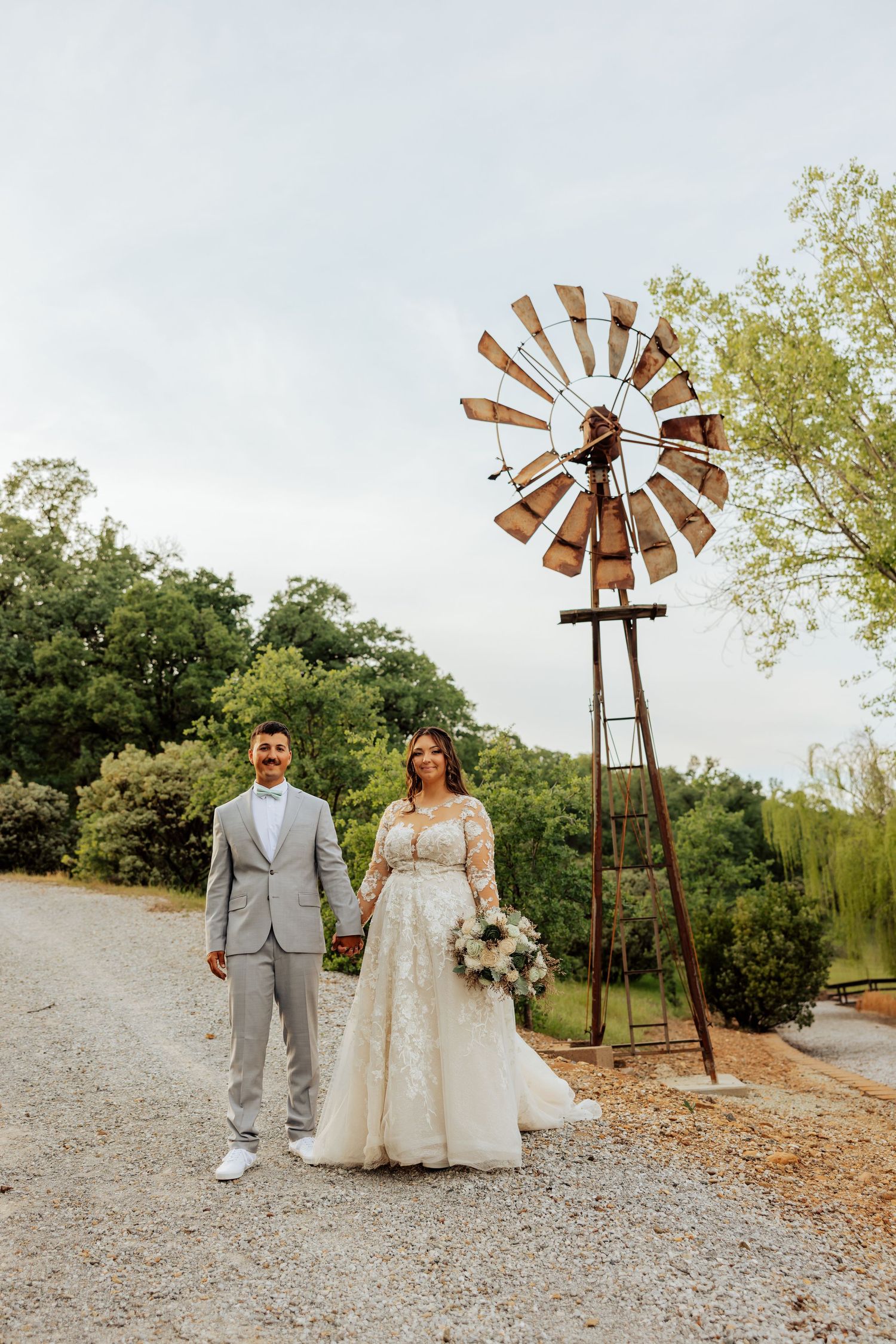 Bride and groom holding hands near a rustic windmill on a gravel path surrounded by lush green trees.