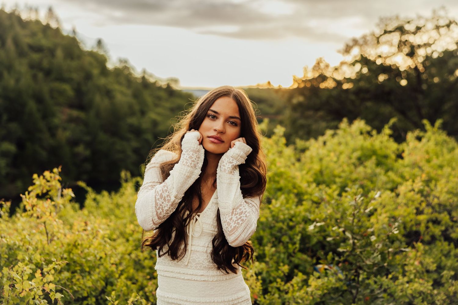 A person in white poses among yellow wildflowers during sunset with mountains in the background.