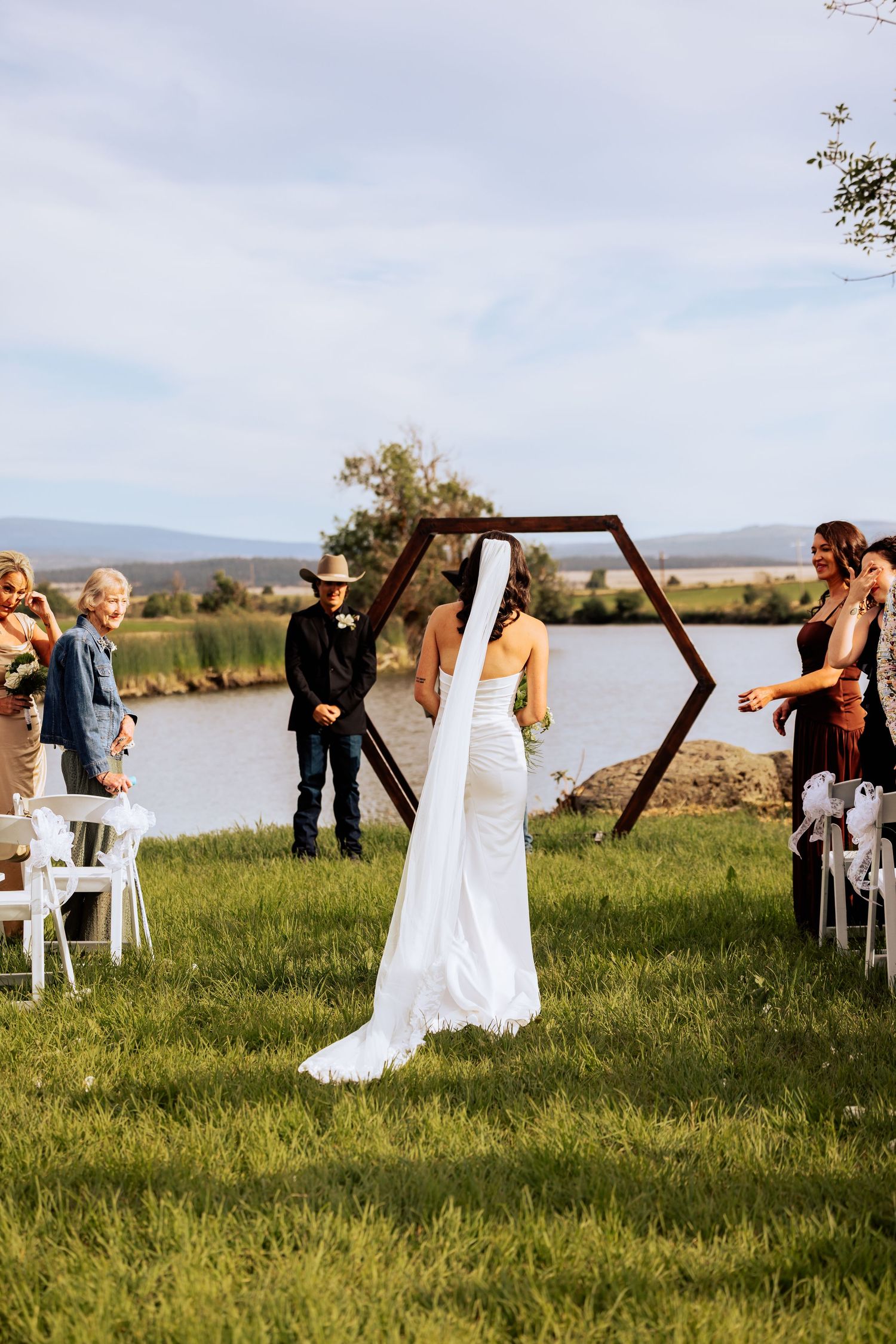 Bride in white gown and veil walks toward hexagonal arch at outdoor lakeside wedding ceremony.