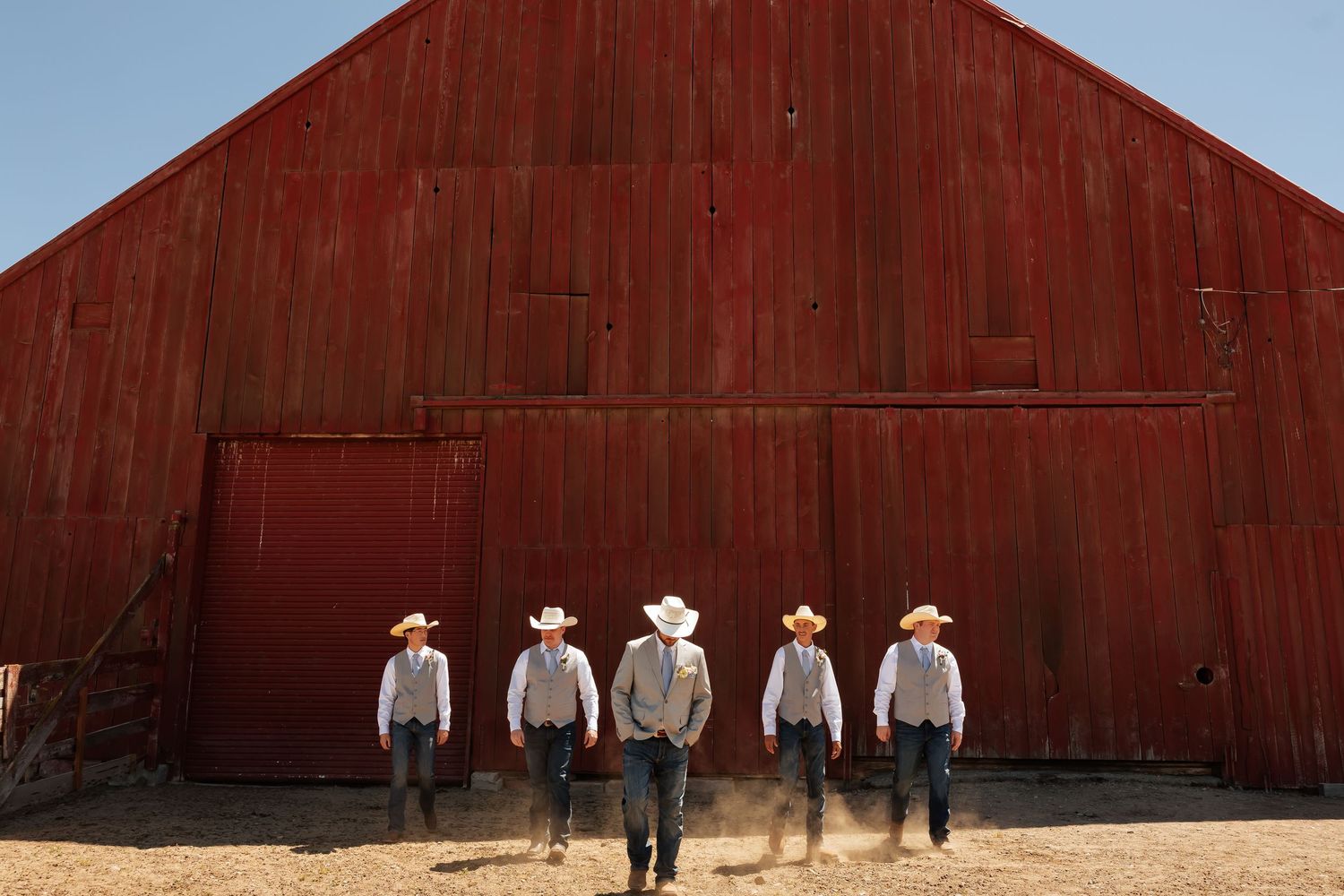 Five cowboys in white shirts and cowboy hats walk away from a rustic red barn on a sunny day.