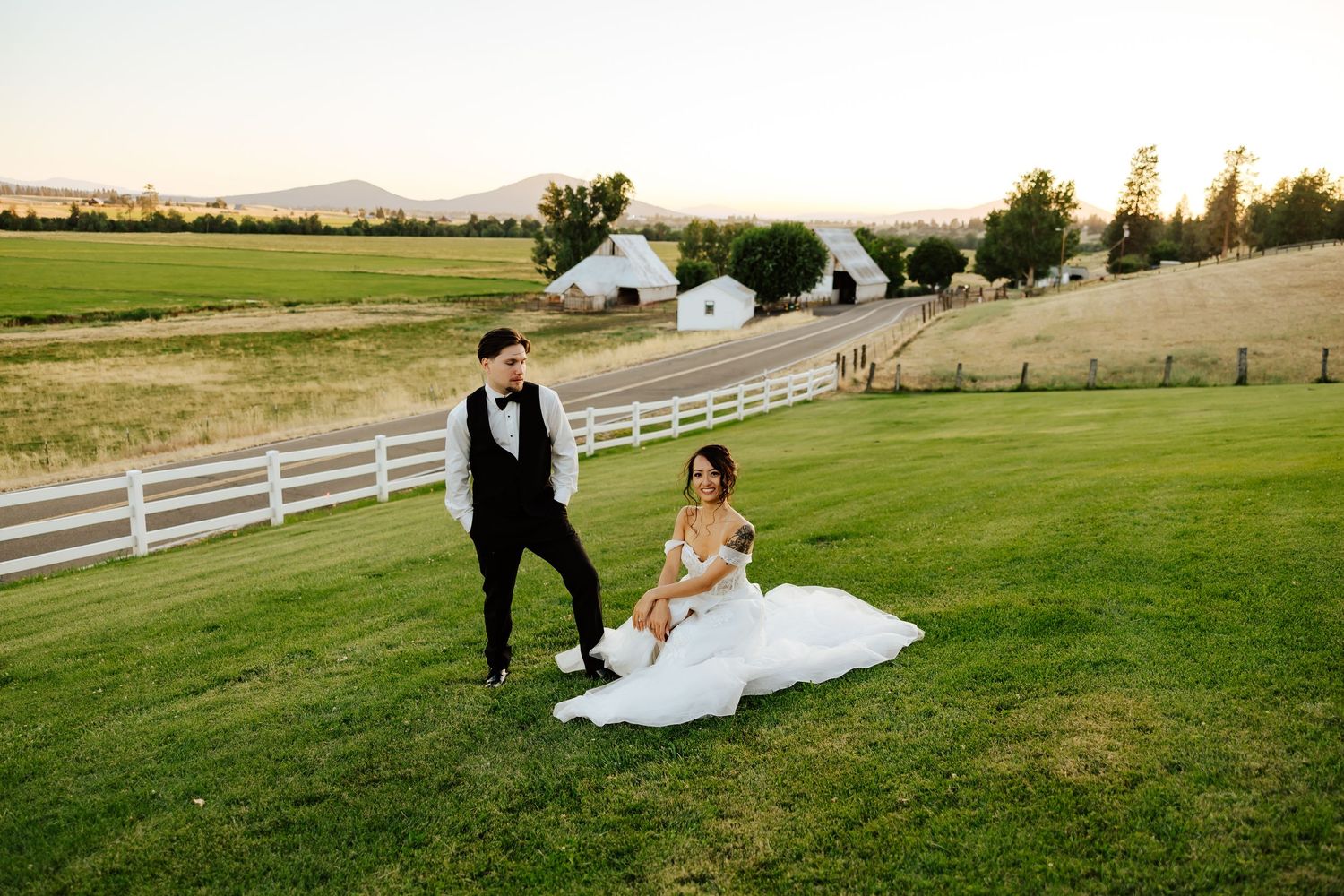 Bride in white gown sits on green lawn while groom stands beside her at a rustic farm wedding venue at sunset.