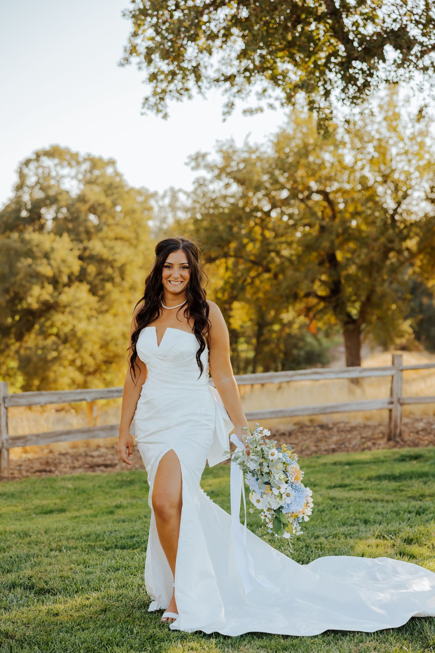 Bride in flowing white dress poses elegantly in outdoor bridal portraits with natural backdrop.