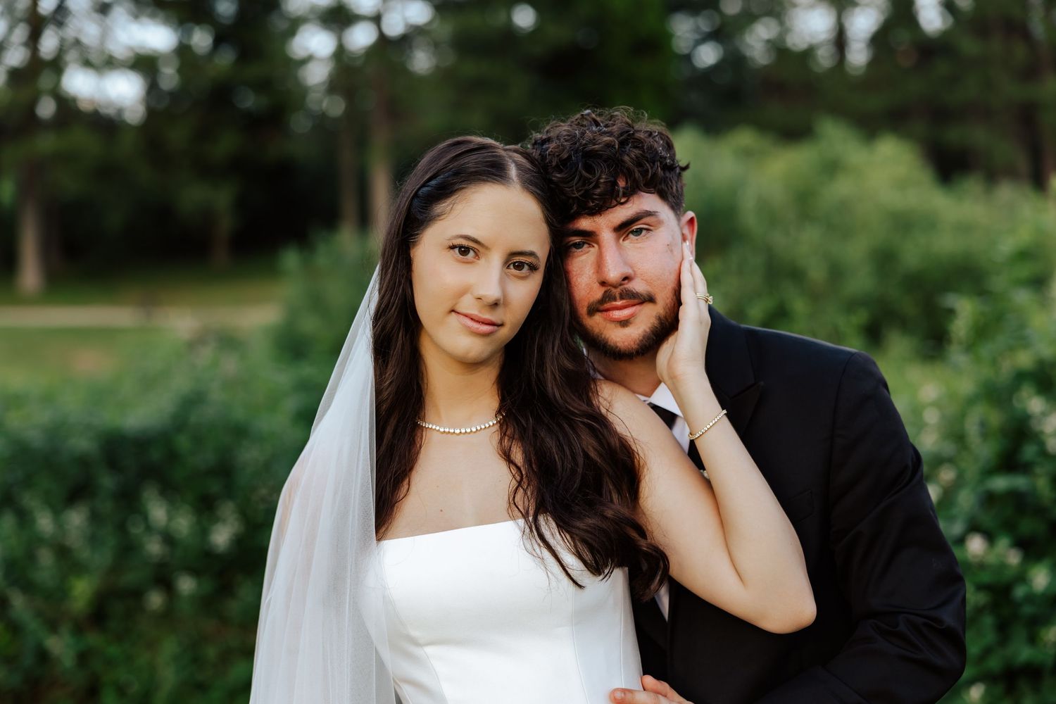 Bride in white strapless gown and veil poses with groom in black suit outdoors with lush green trees behind them.