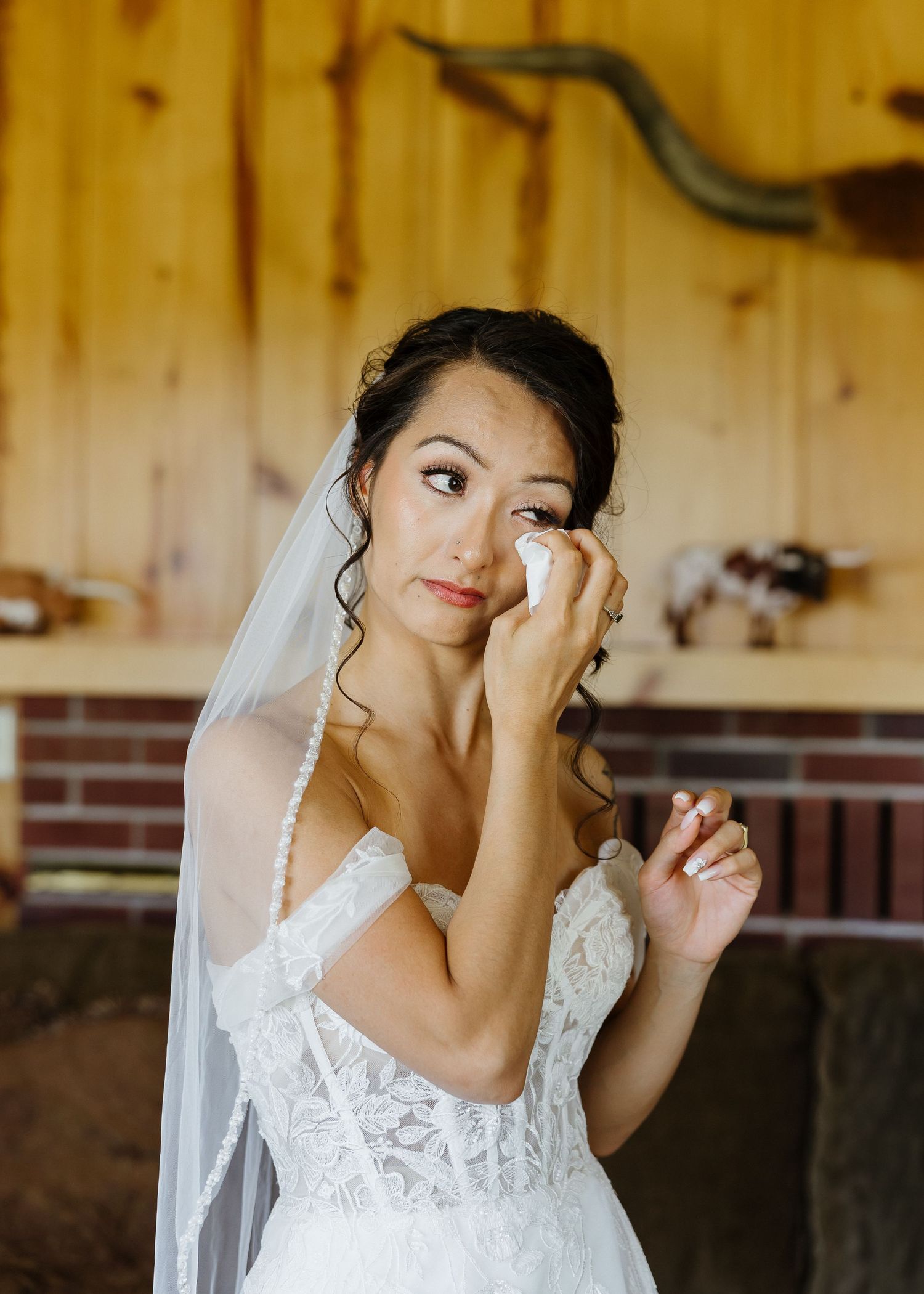 Emotional bride in lace wedding dress and veil dabbing tears with tissue in rustic barn setting.