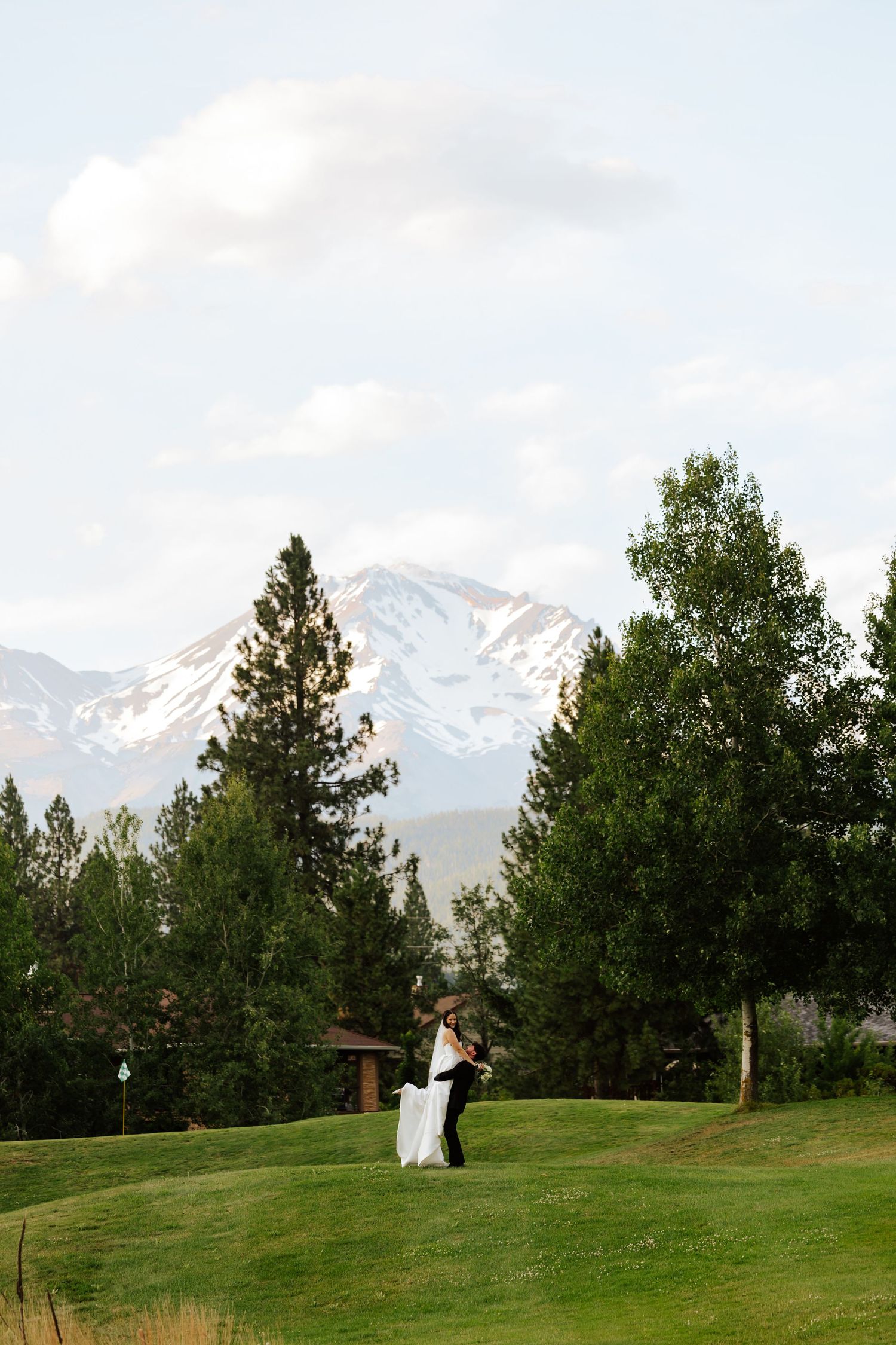 Bride and groom share romantic embrace on golf course with stunning snow-capped mountain backdrop.