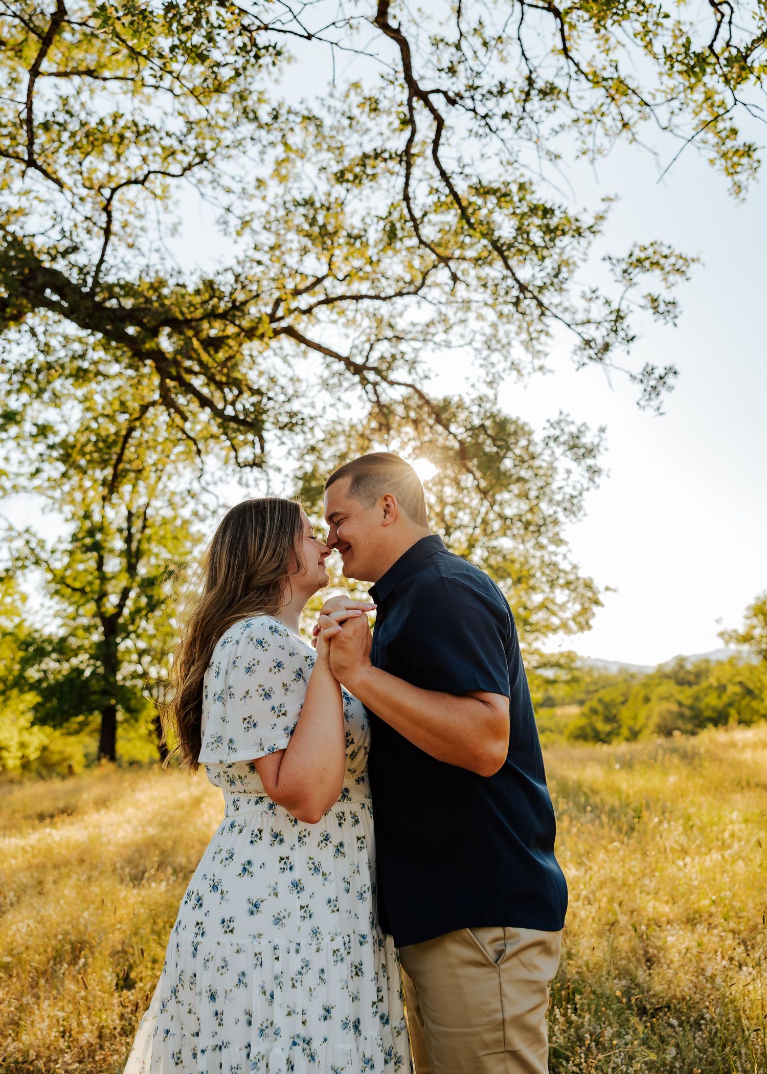 Couple sharing romantic moment under oak tree in sunlit meadow, woman in floral dress holding hands.