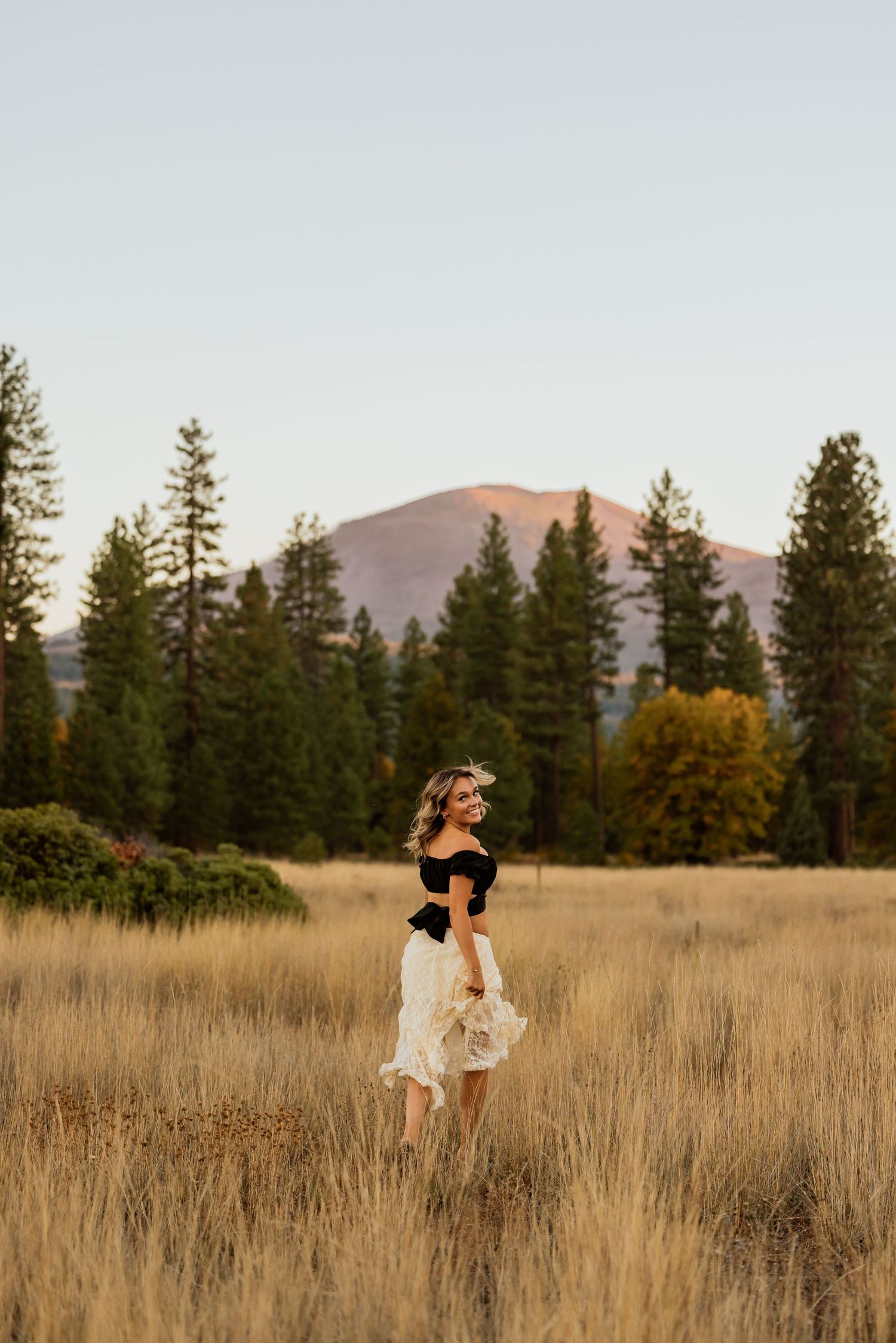 Artistic portrait in tall grass with mountain landscape and evergreen forest backdrop.