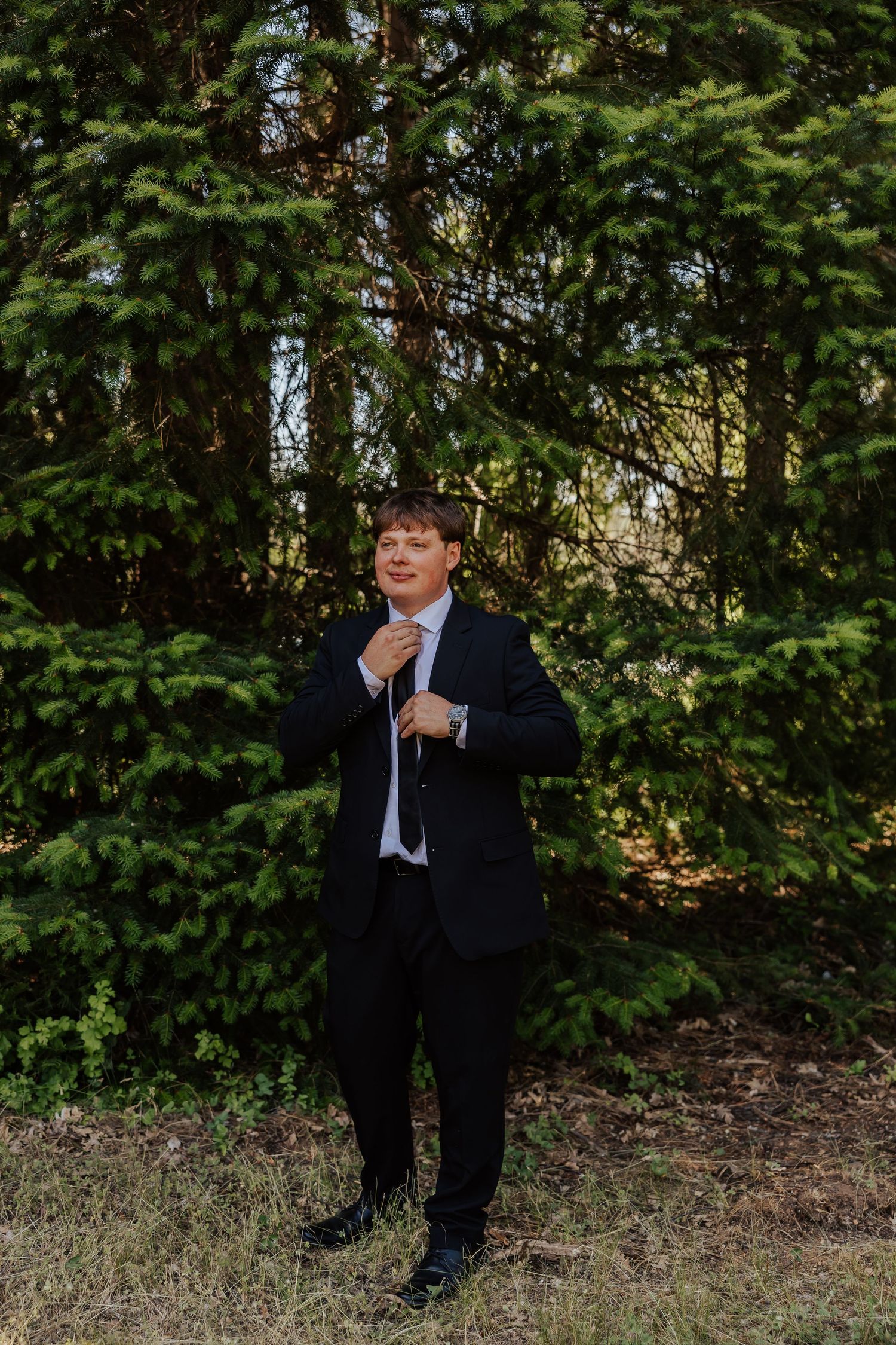 Dapper groom in black suit adjusts his tie while standing in front of lush green pine trees outdoors.
