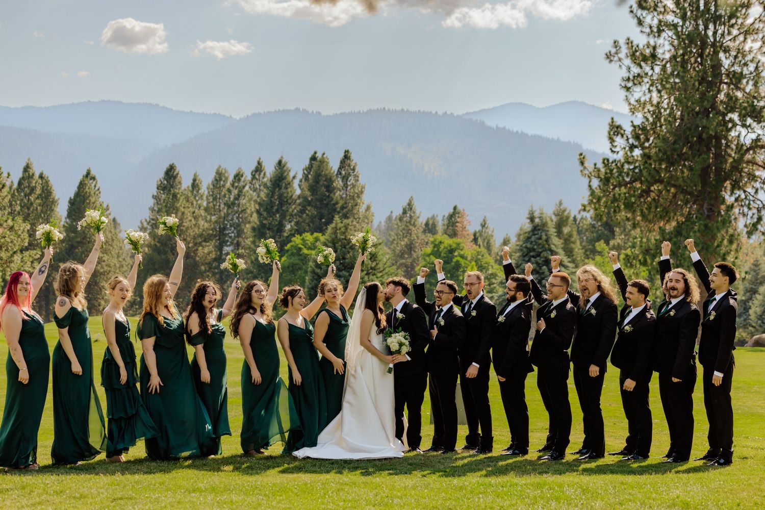 Wedding party celebrating outdoors with mountain backdrop, bridesmaids in emerald gowns and groomsmen in black suits raising bouquets.