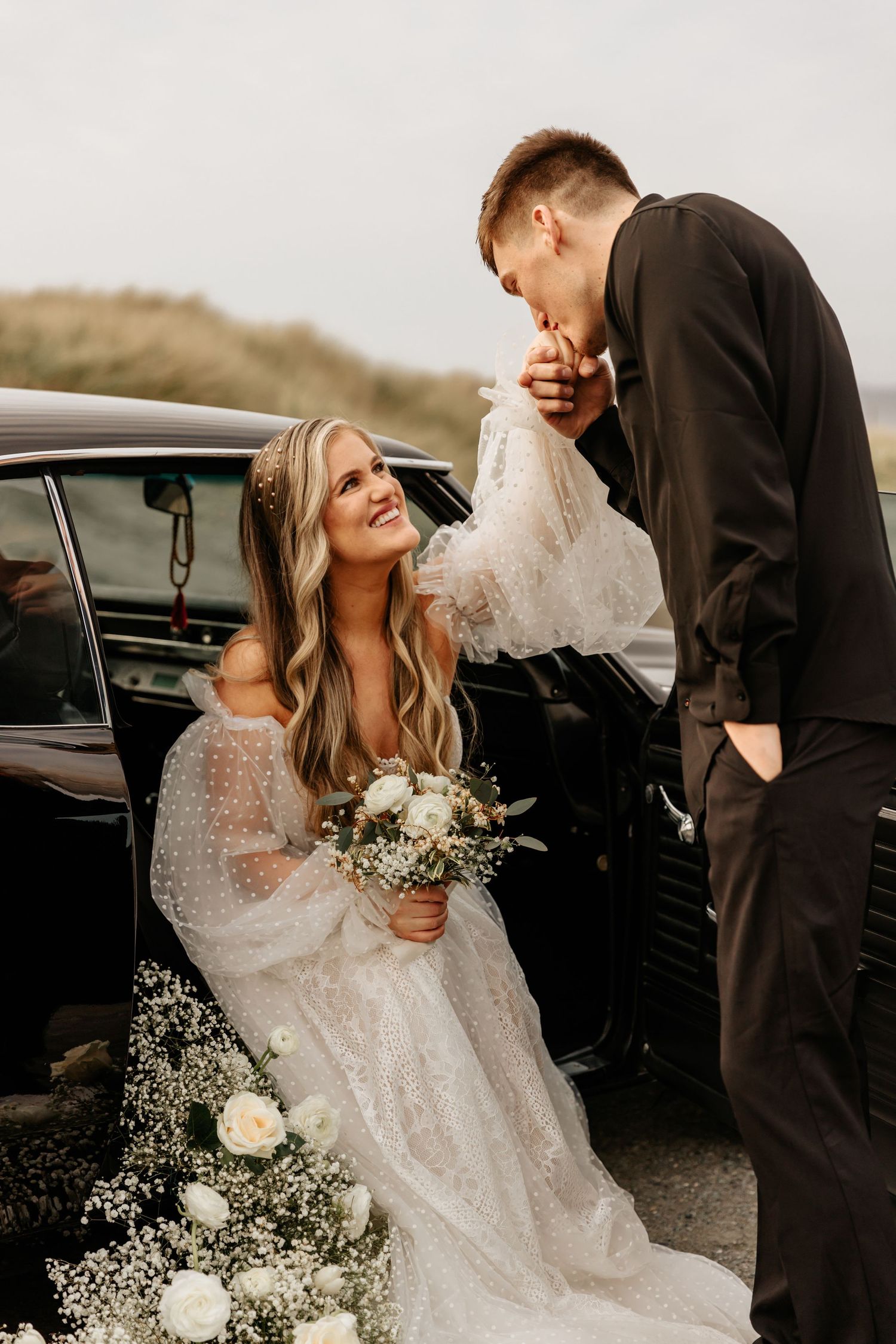Sweet moment between newlyweds by classic car with bride holding white rose bouquet.