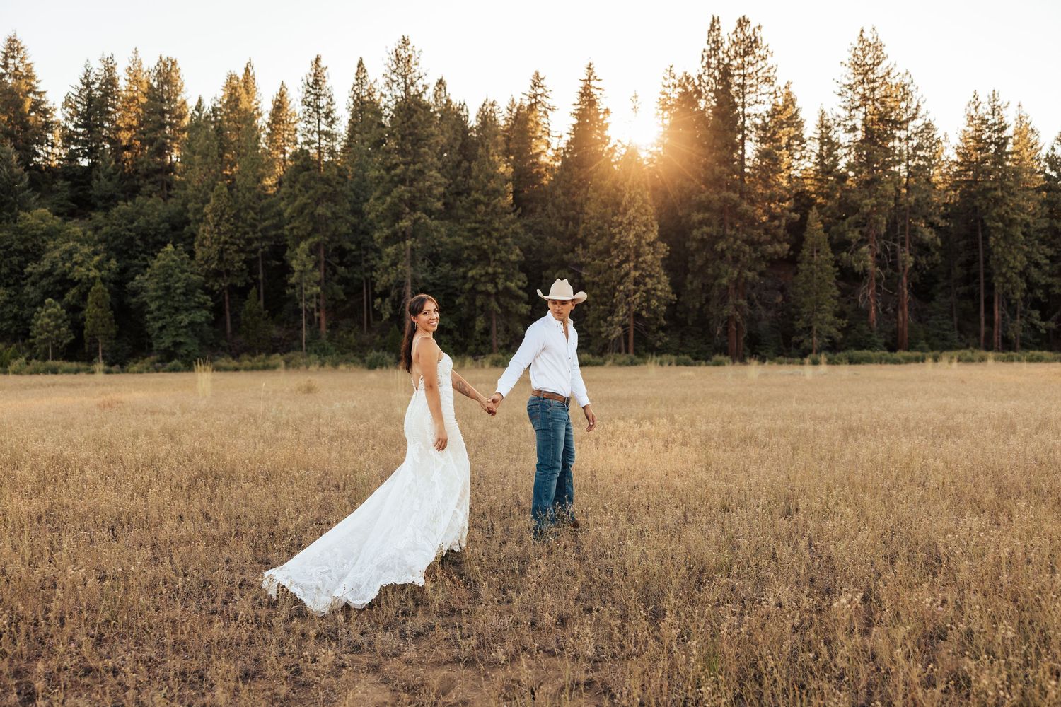 A romantic sunset photo of newlyweds walking hand in hand through a golden field with pine trees.
