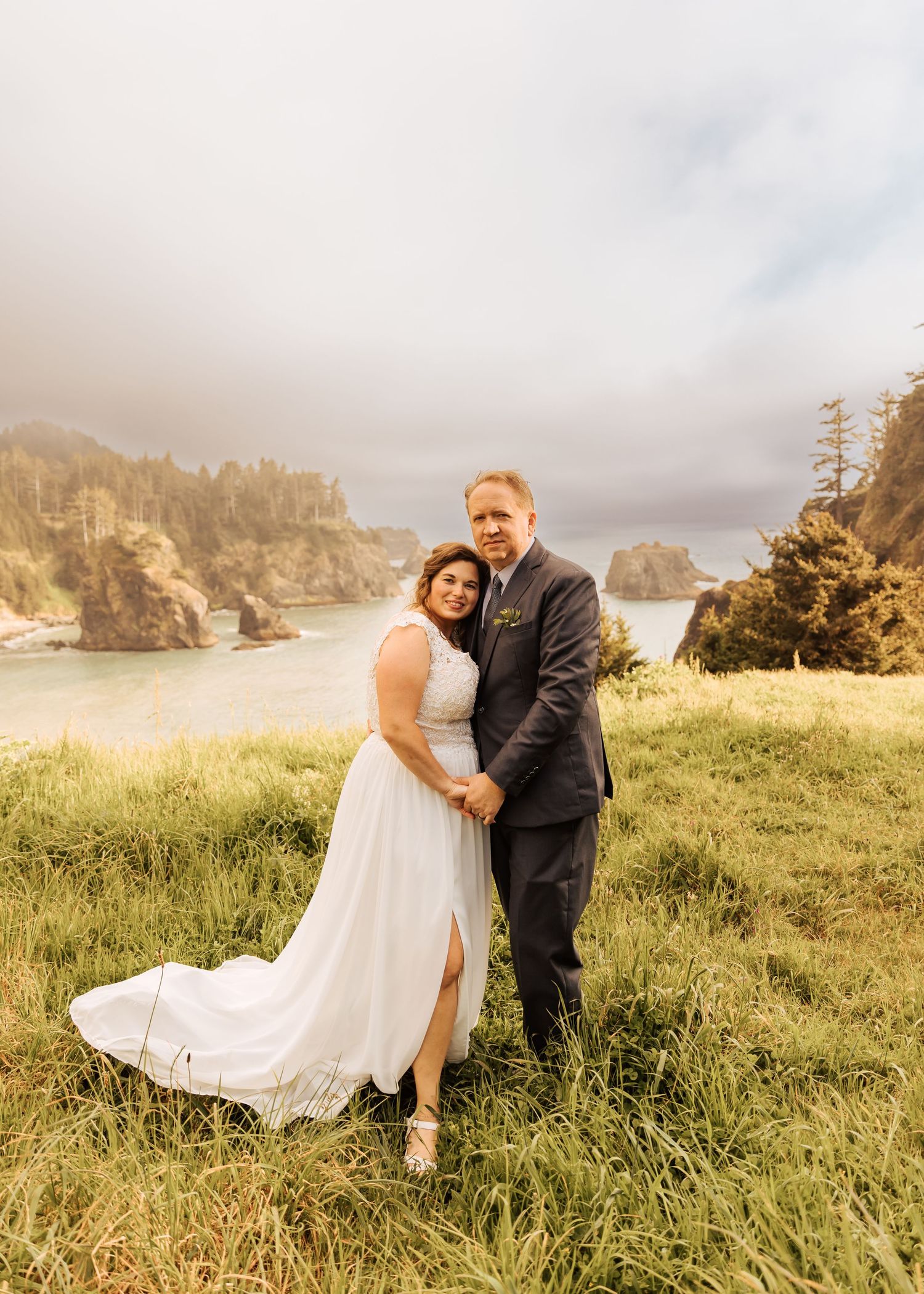 A romantic wedding photoshoot on a grassy coastal cliff overlooking the ocean at sunset.