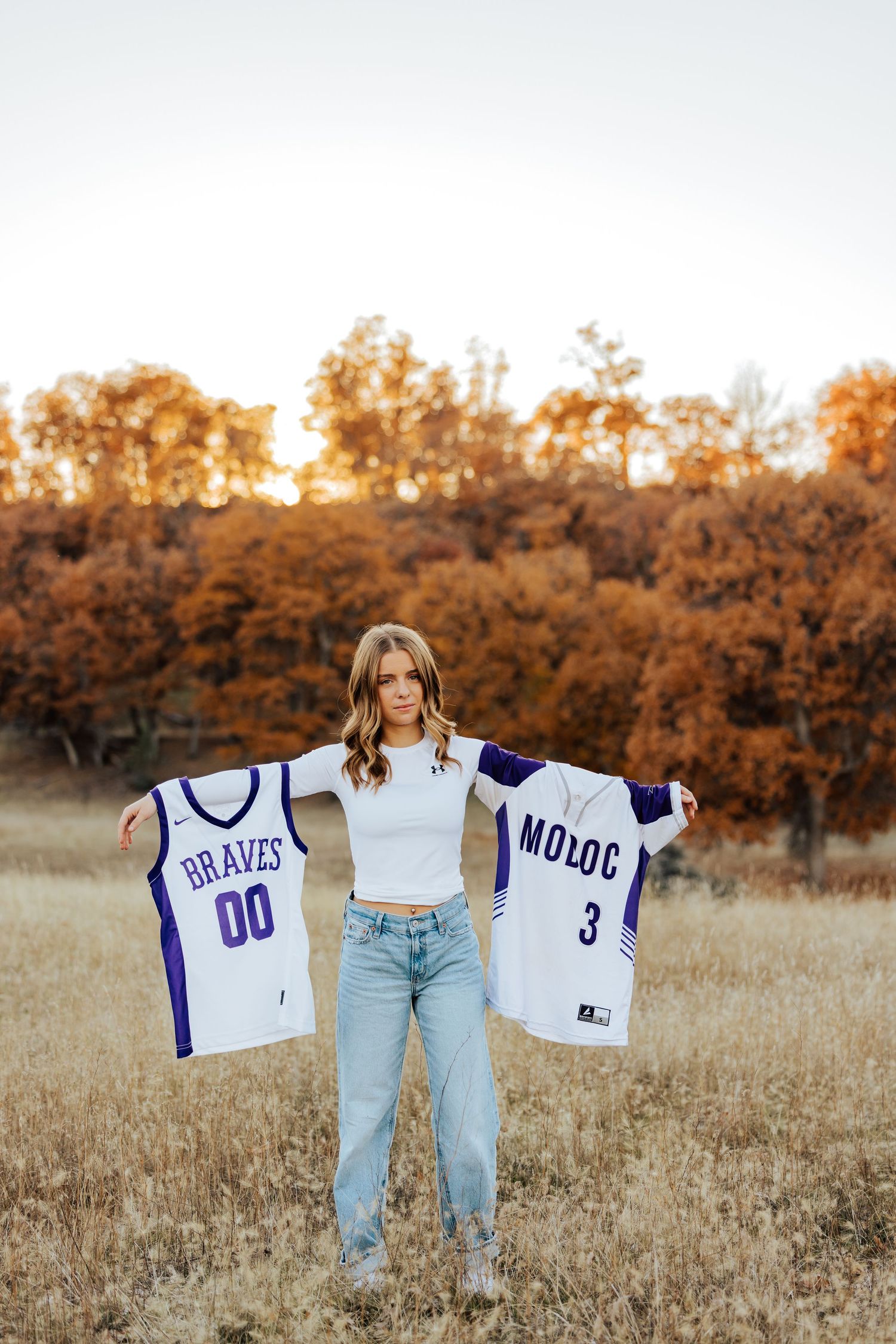 Senior displaying sports jerseys during golden hour photoshoot with fall colors.