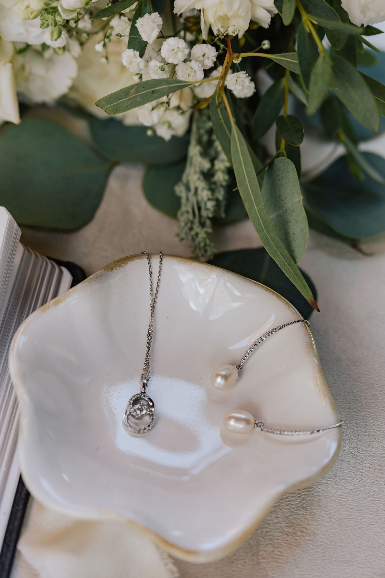 Silver necklace and pearl bracelet displayed in a white ceramic dish with elegant floral arrangement backdrop.