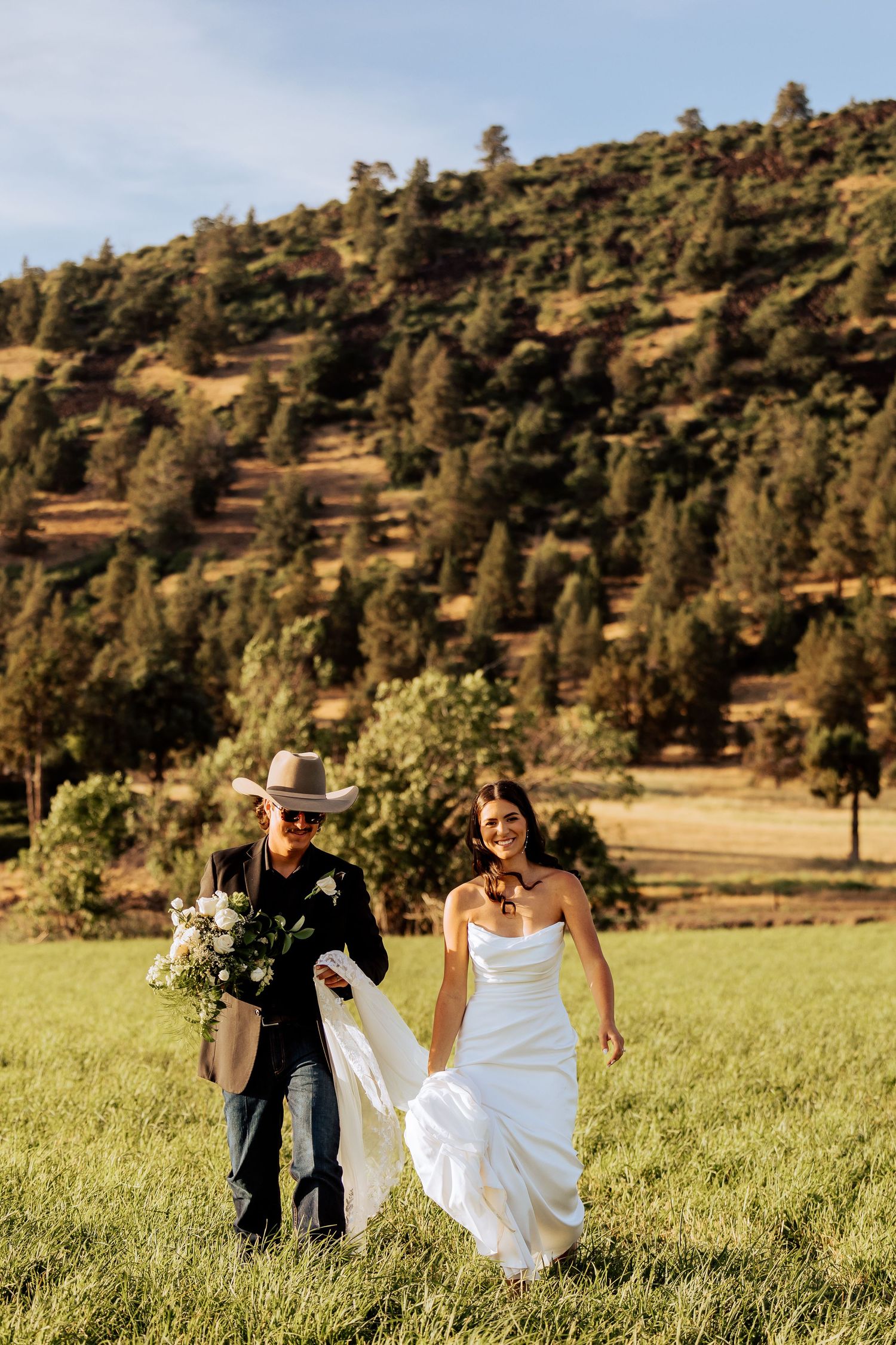 A bride in a white gown and groom in a cowboy hat walk through a sunlit green field with a forested hill backdrop.