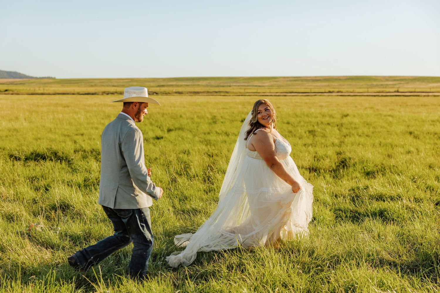 Bride in white gown and groom in gray suit walk through green field, bride holding veil train on sunny day.