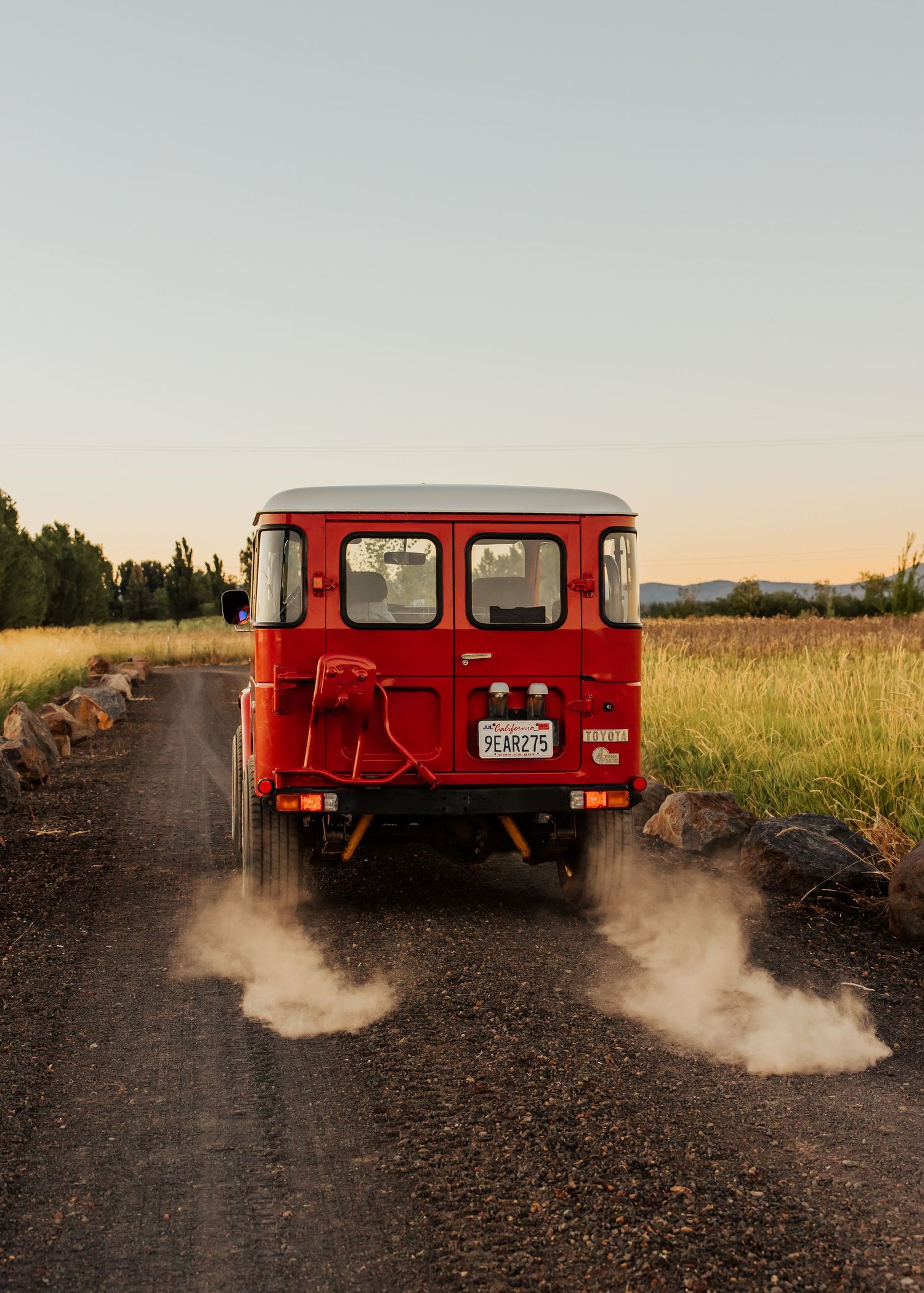 A red Toyota Land Cruiser kicks up dust on a dirt road at sunset.