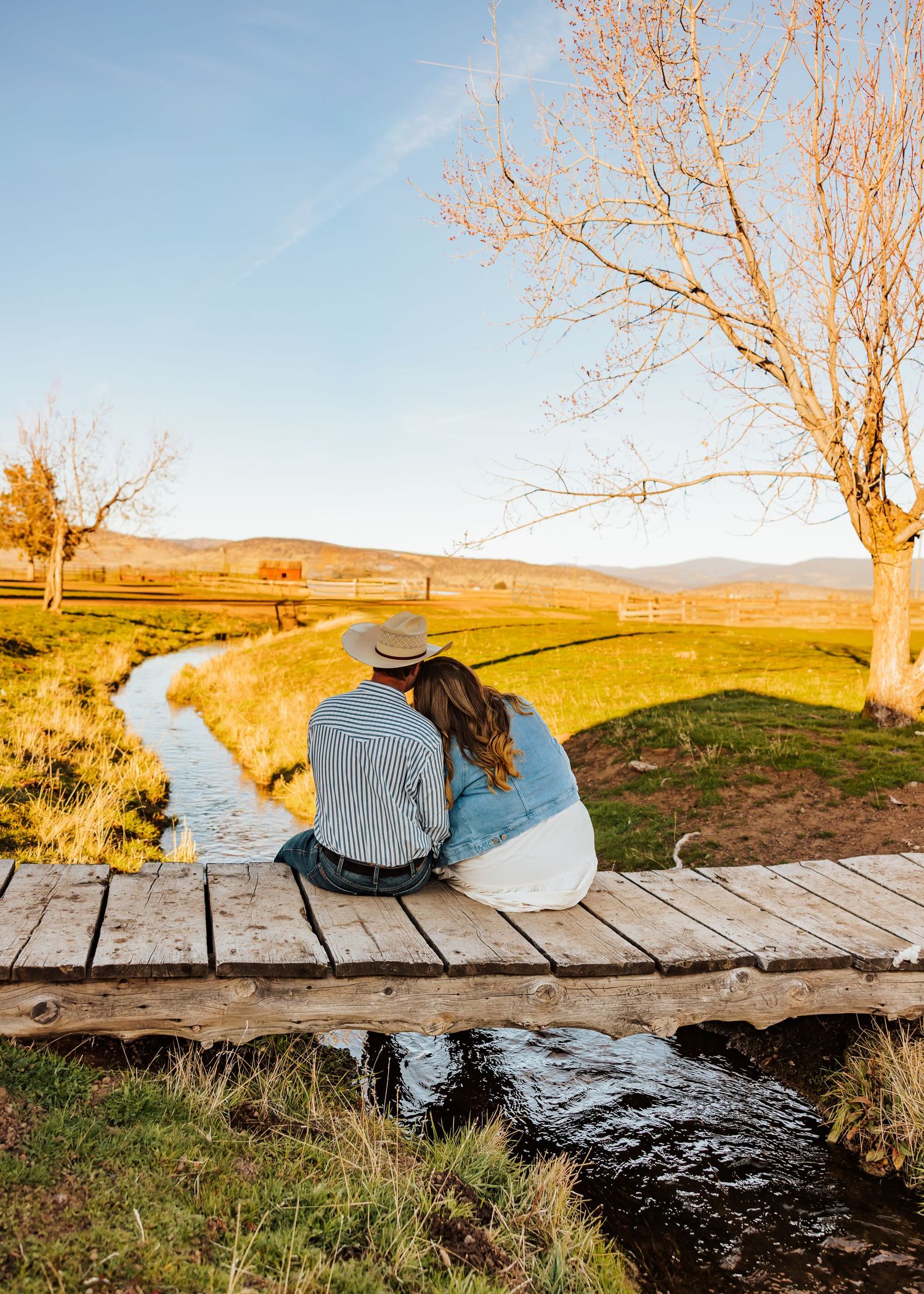 A couple sits together on wooden bridge overlooking scenic creek and rolling hills.
