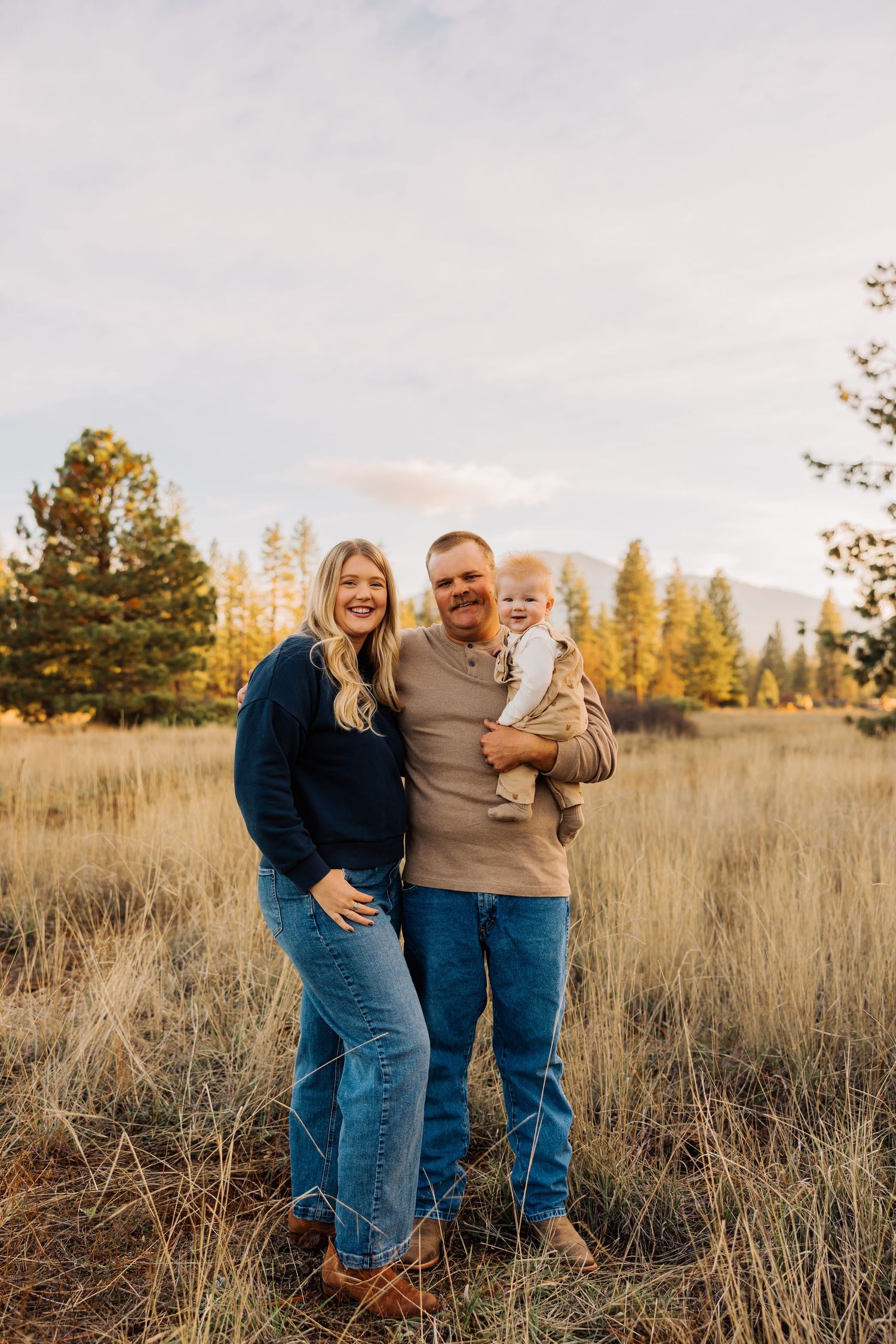 Happy family portrait in rustic field setting with mountains and sunset lighting.