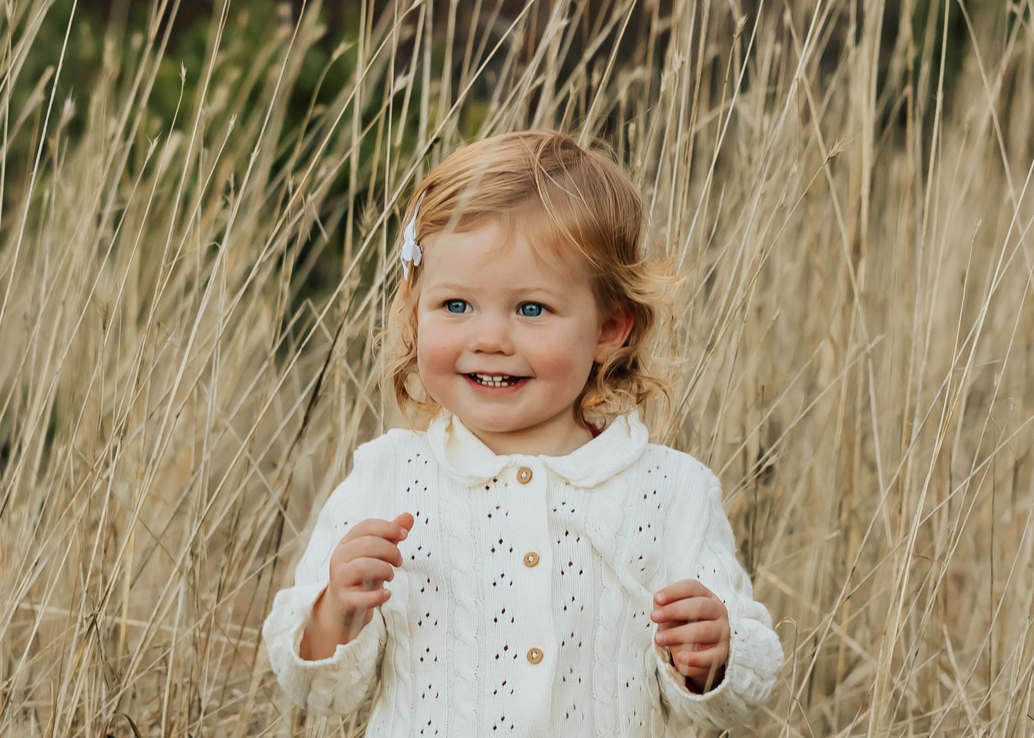 Toddler in white sweater smiles while playing in tall autumn grass.