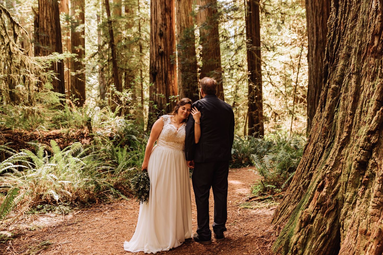 A couple shares intimate moments along a serene redwood forest path during their wedding day.