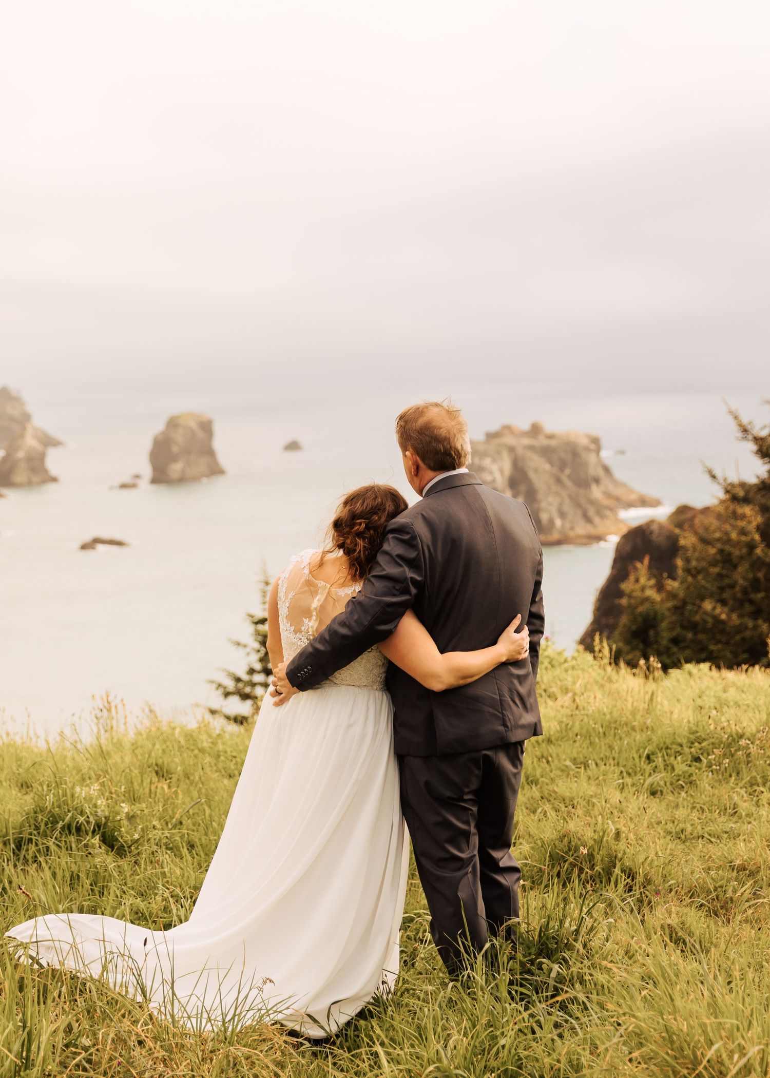 A couple shares tender moments on a scenic coastal hillside with ocean rock formations in the background.