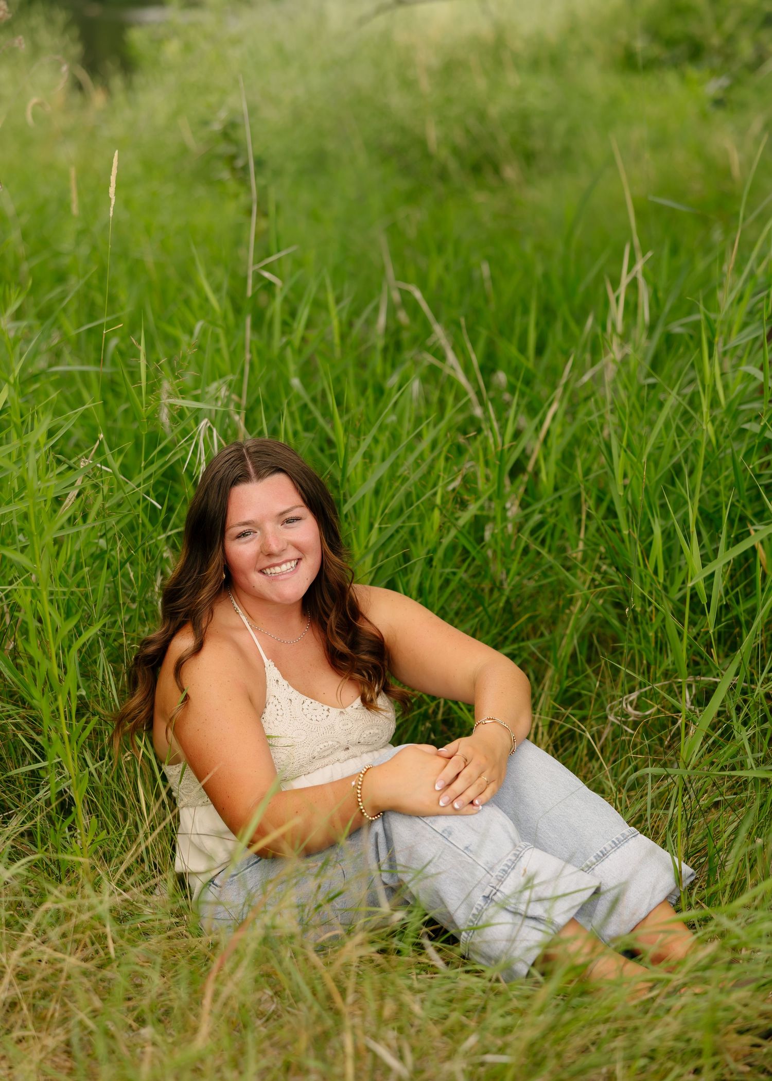 Smiling young woman sitting in tall green grass wearing a white lace top and light blue jeans.