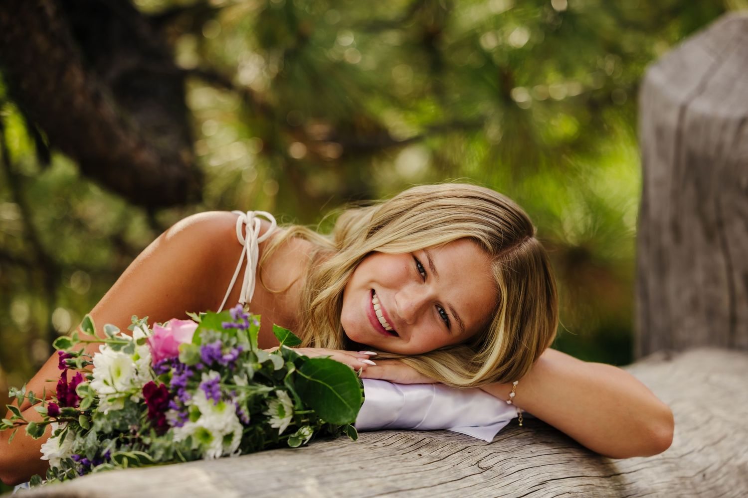 Smiling bride resting on rustic wooden surface holding colorful wedding bouquet with purple and white flowers outdoors.