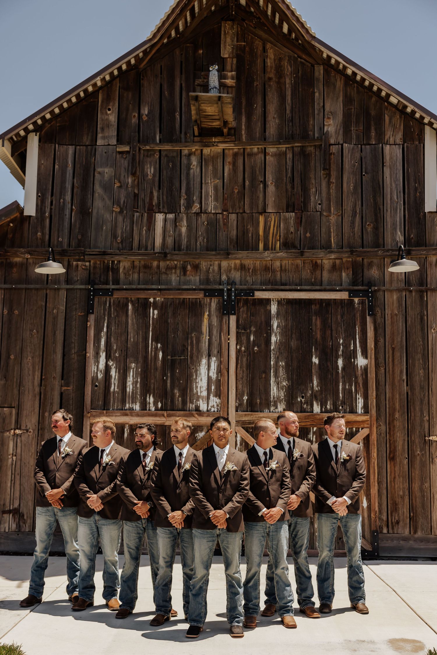 Groom and groomsmen in brown suits and jeans posing in front of a rustic wooden barn at a country wedding.