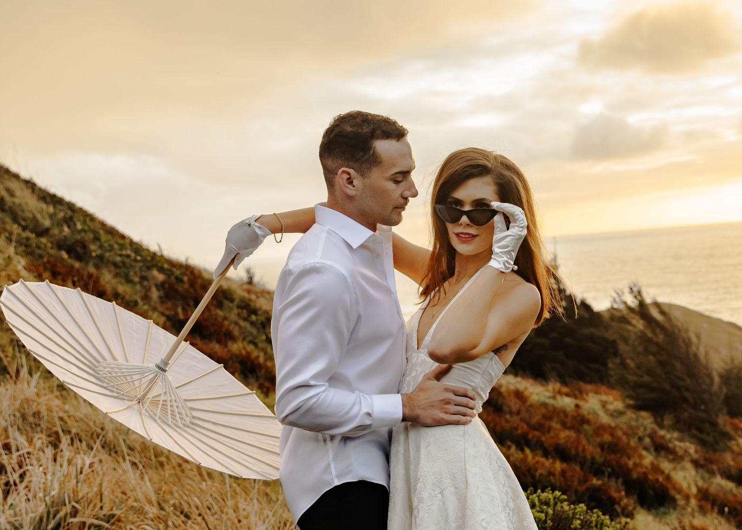 Couple in formal attire posing together at sunset on a coastal hillside.