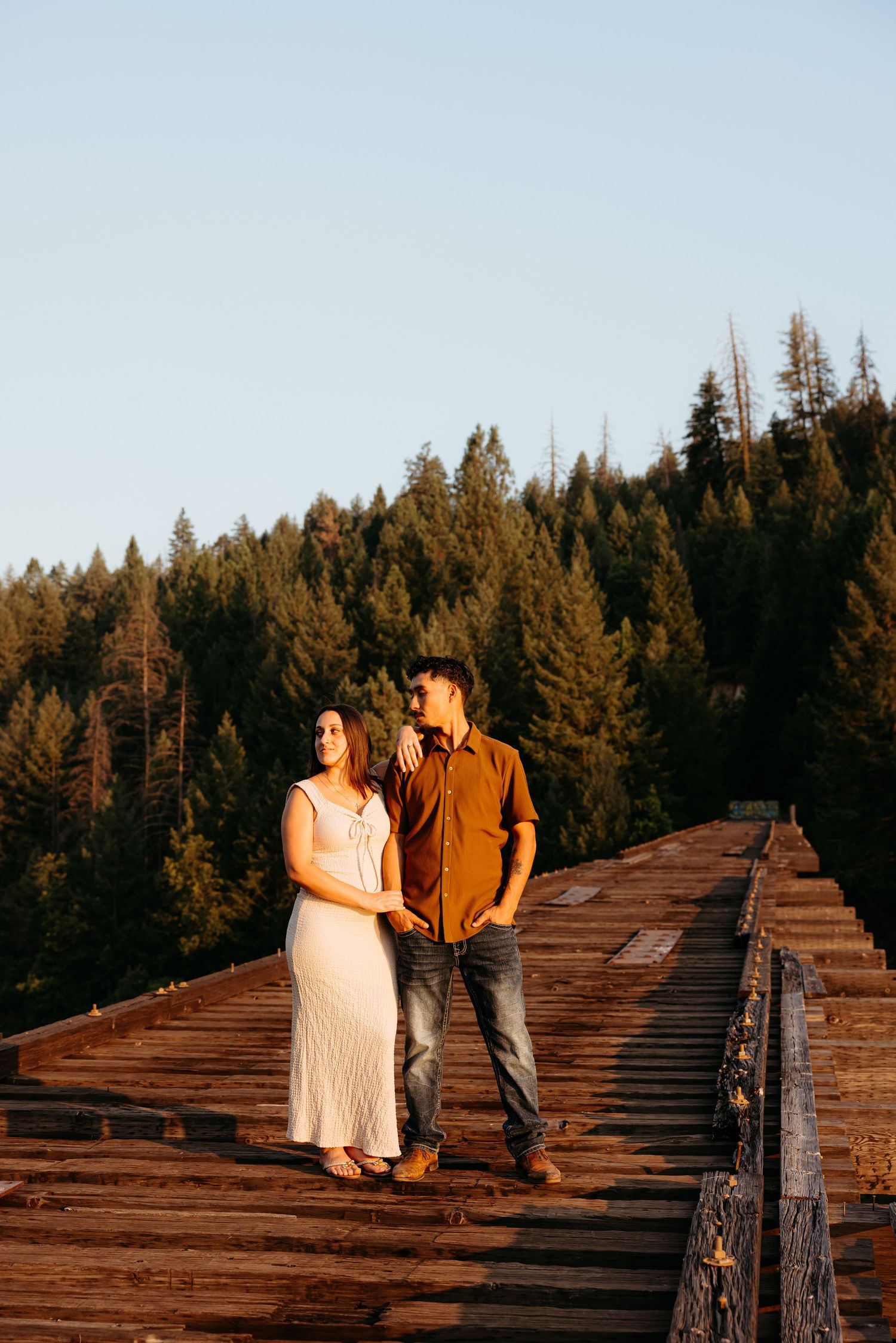 Couple stands together on rustic wooden bridge during golden hour in forest.